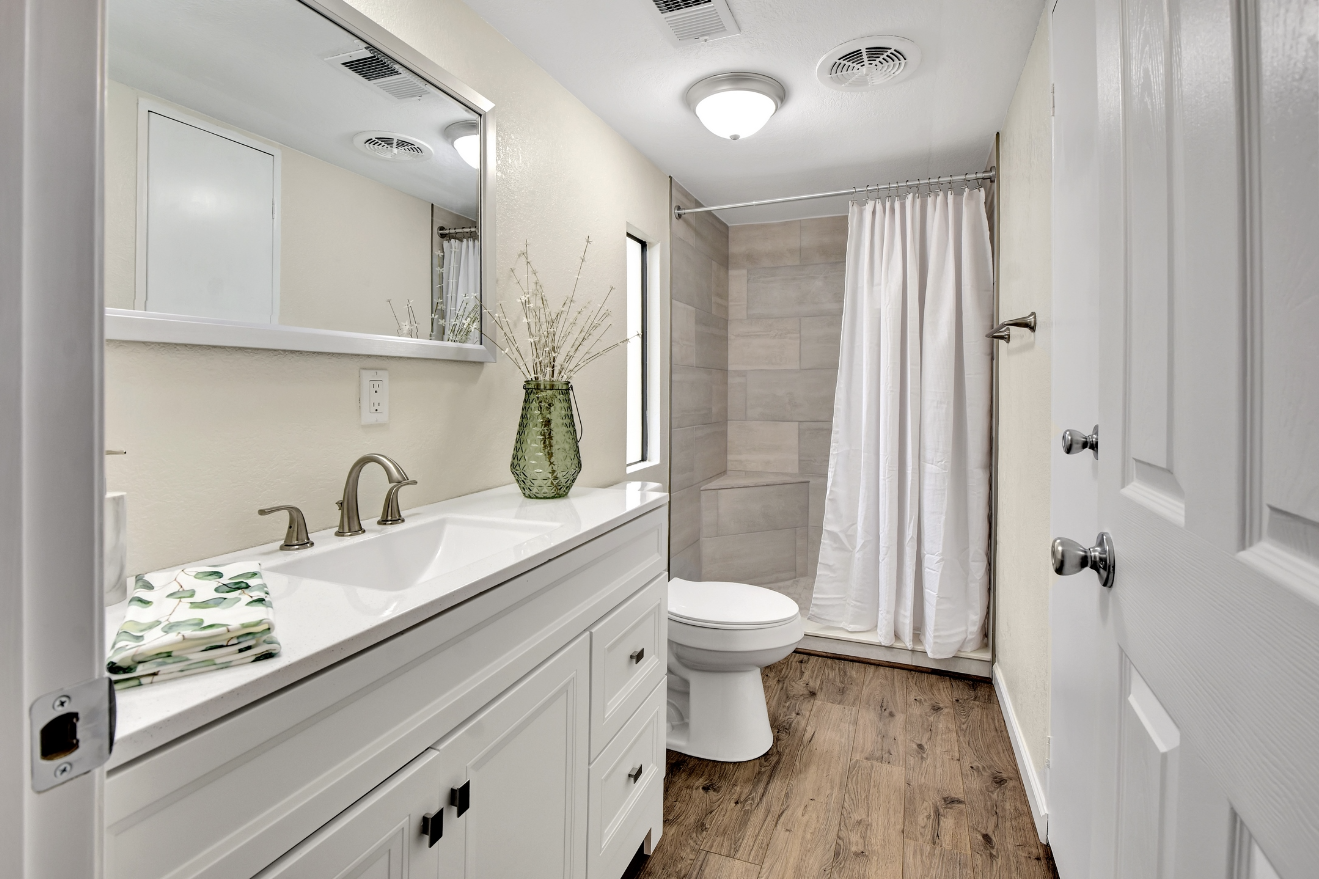 A clean bathroom with a white vanity, a mirror, a sink with a brushed nickel faucet, a green vase with dried branches, a folded towel with a leaf pattern, a toilet, a small window, and a shower with a white curtain and beige tile walls. The floor is 