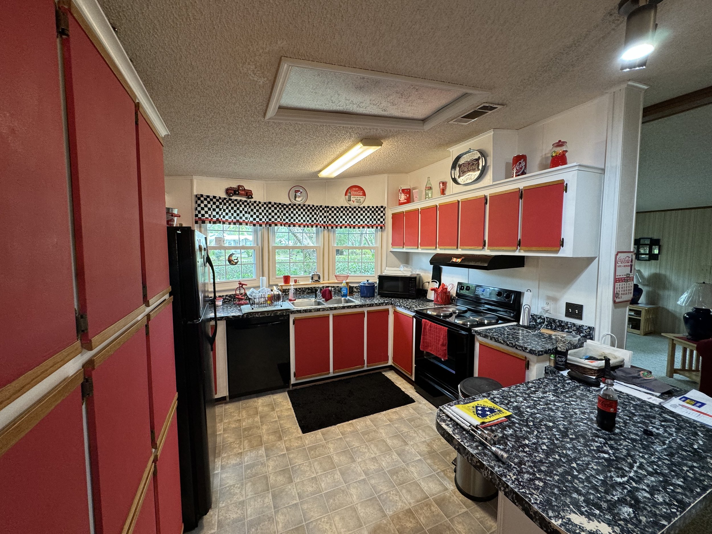 A cozy kitchen with red cabinets, black appliances, granite countertops, and a window with checkered valance. Decor includes vintage-themed items and Coca-Cola collectibles.