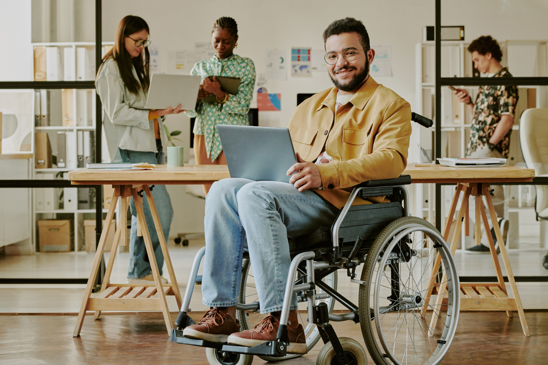 An individual in a wheelchair works on a laptop that is seated on their lap. They are smiling at the camera while their coworkers in the background are standing around the office, looking at shared devices.