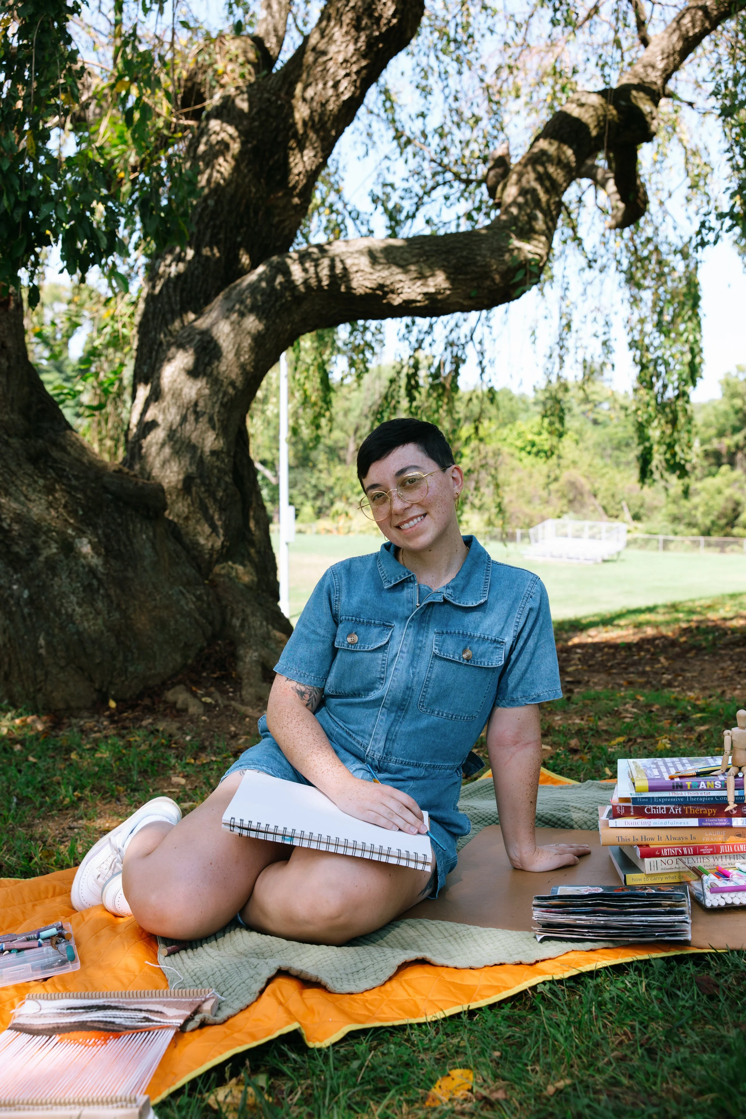 Nonbinary art therapist Kristy Leone sitting outdoors in Towson Maryland holding oil pastels and a blank canvas before an art therapy session.