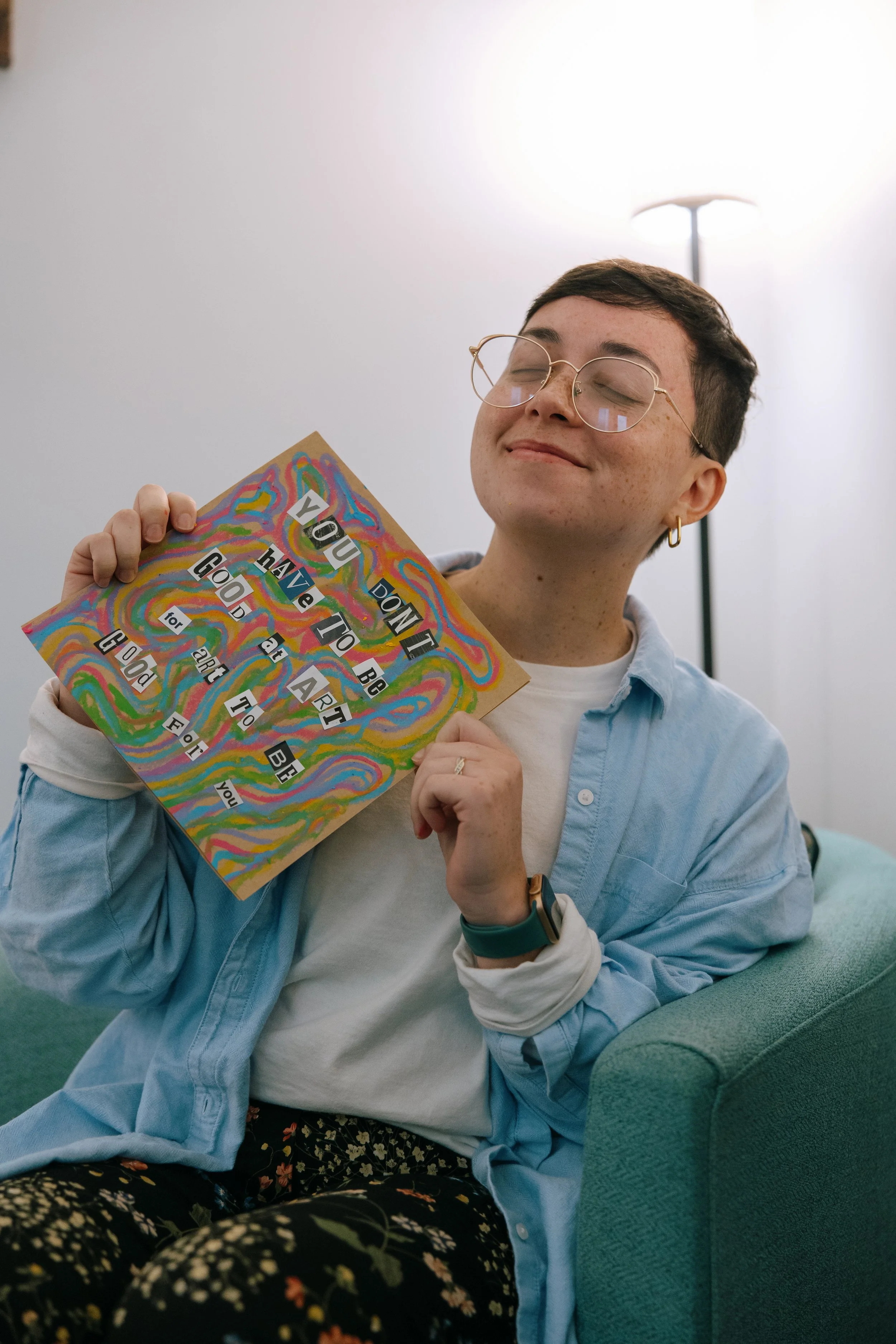 Nonbinary art therapist in Maryland, Kristy Leone, holding an oil pastel picture that says you don't have to be good at art for art to be good for you in their office in Timonium Maryland.