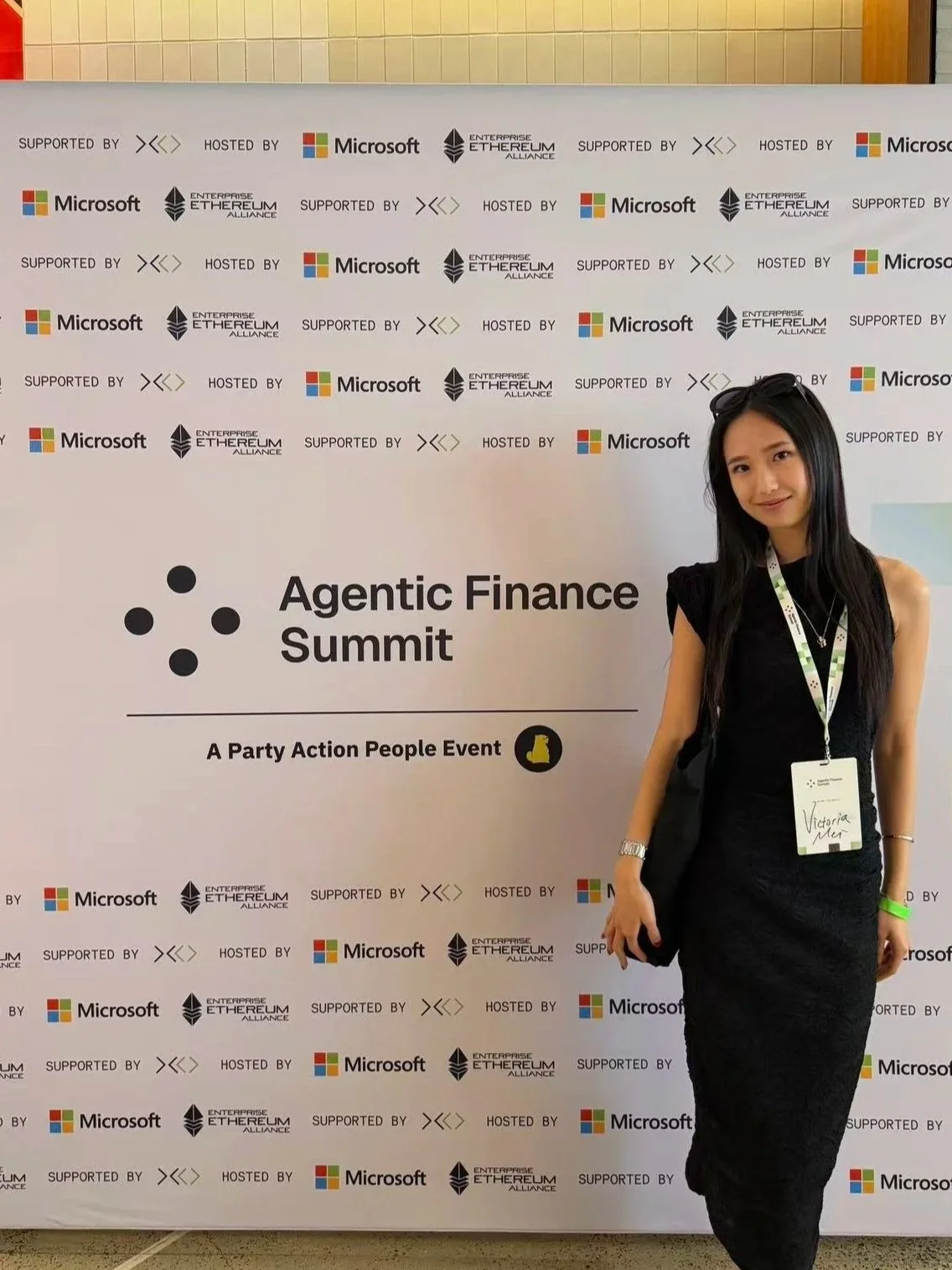 A woman wearing a black dress and a name tag standing in front of a backdrop with logos of Microsoft, Ethereum Alliance, and other organizations. The backdrop indicates the event is the 'Agentic Finance Summit,' described as a 'Party Action People Event.'