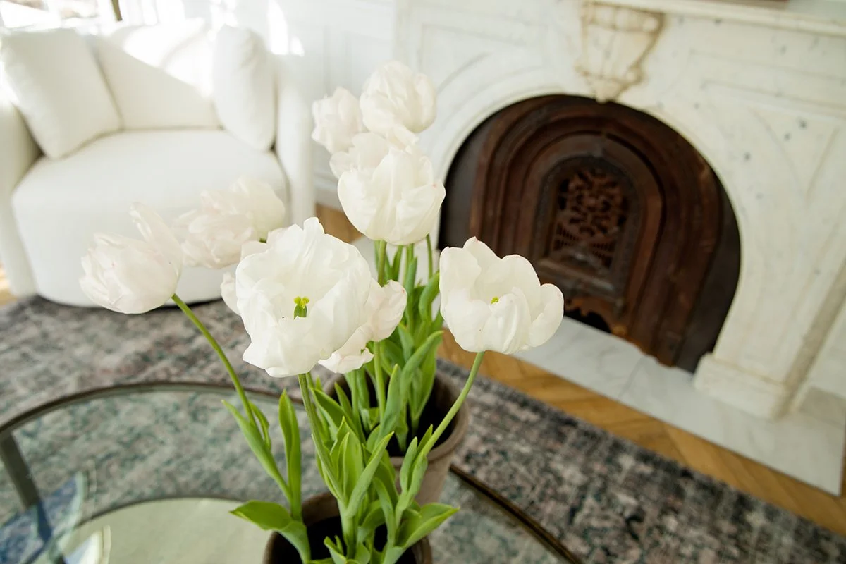 A potted white tulip plant on a glass table in a living room with a white couch, a patterned rug, and a fireplace with a decorative dark metal fire grate.