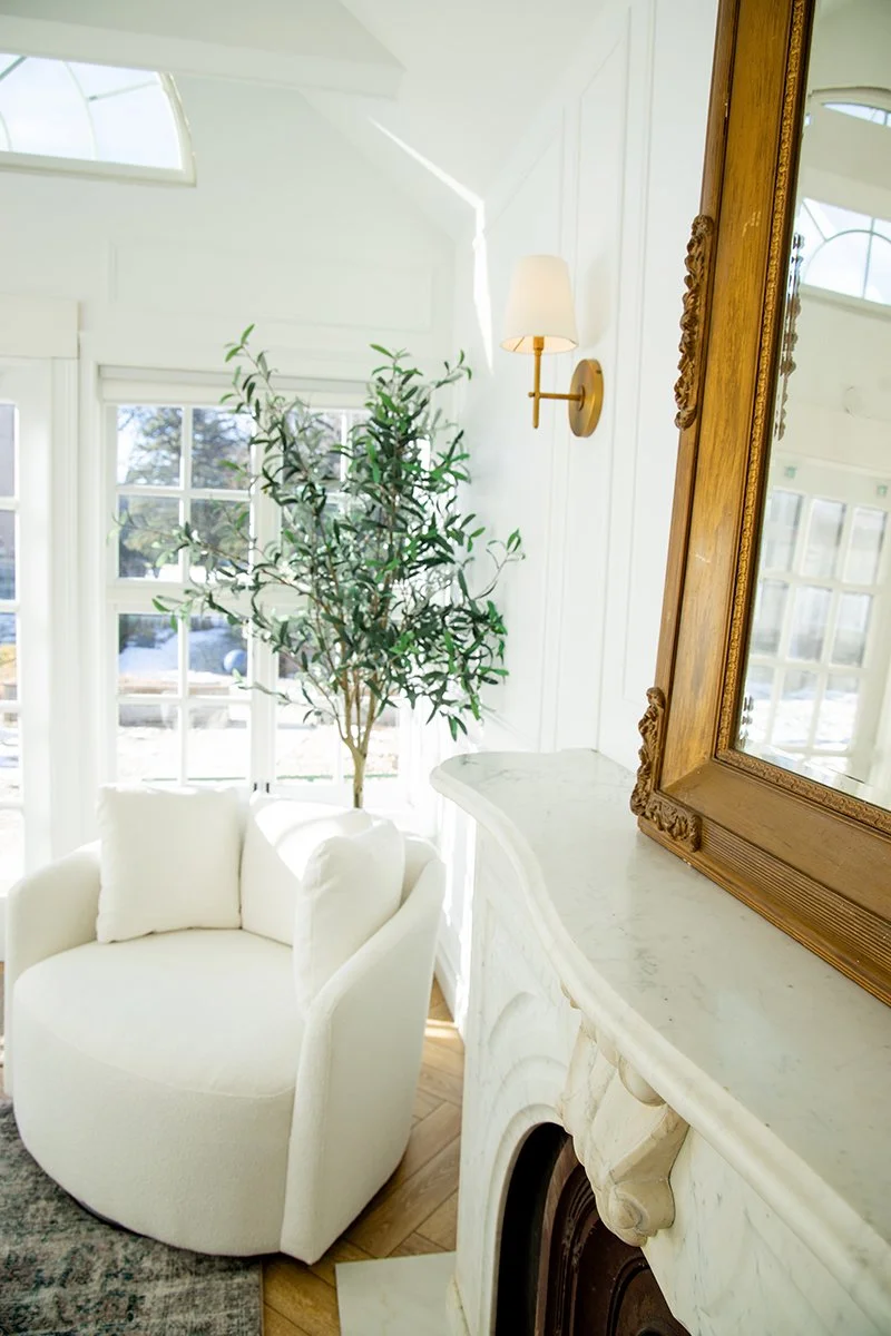 Bright interior corner with a white upholstered armchair, a tall potted plant, a marble fireplace, and a large ornate wooden framed mirror, with a window in the background.