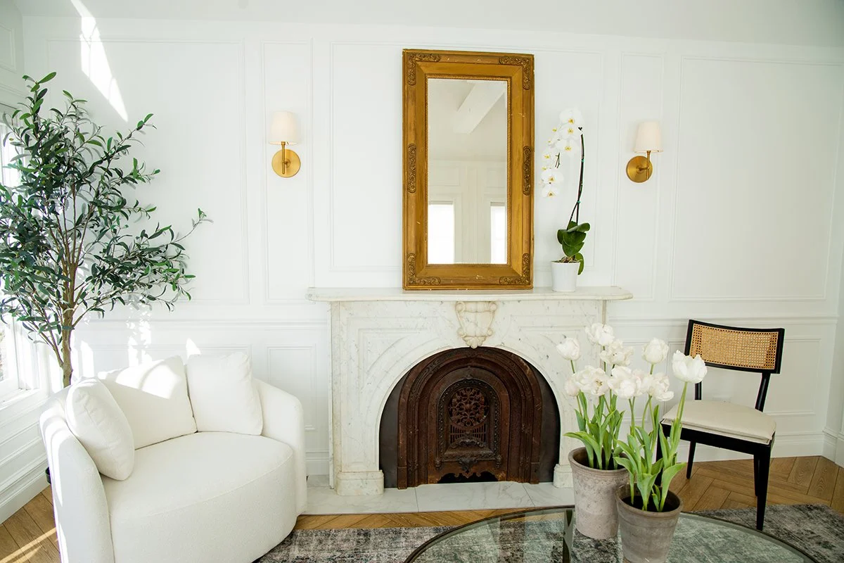 Living room corner with a white sofa, a white marble fireplace with an ornate metal insert, a large gold-framed mirror above the fireplace, two wall-mounted lamps, a potted orchid plant on the mantel, another potted tulip plant on a glass-top table,
