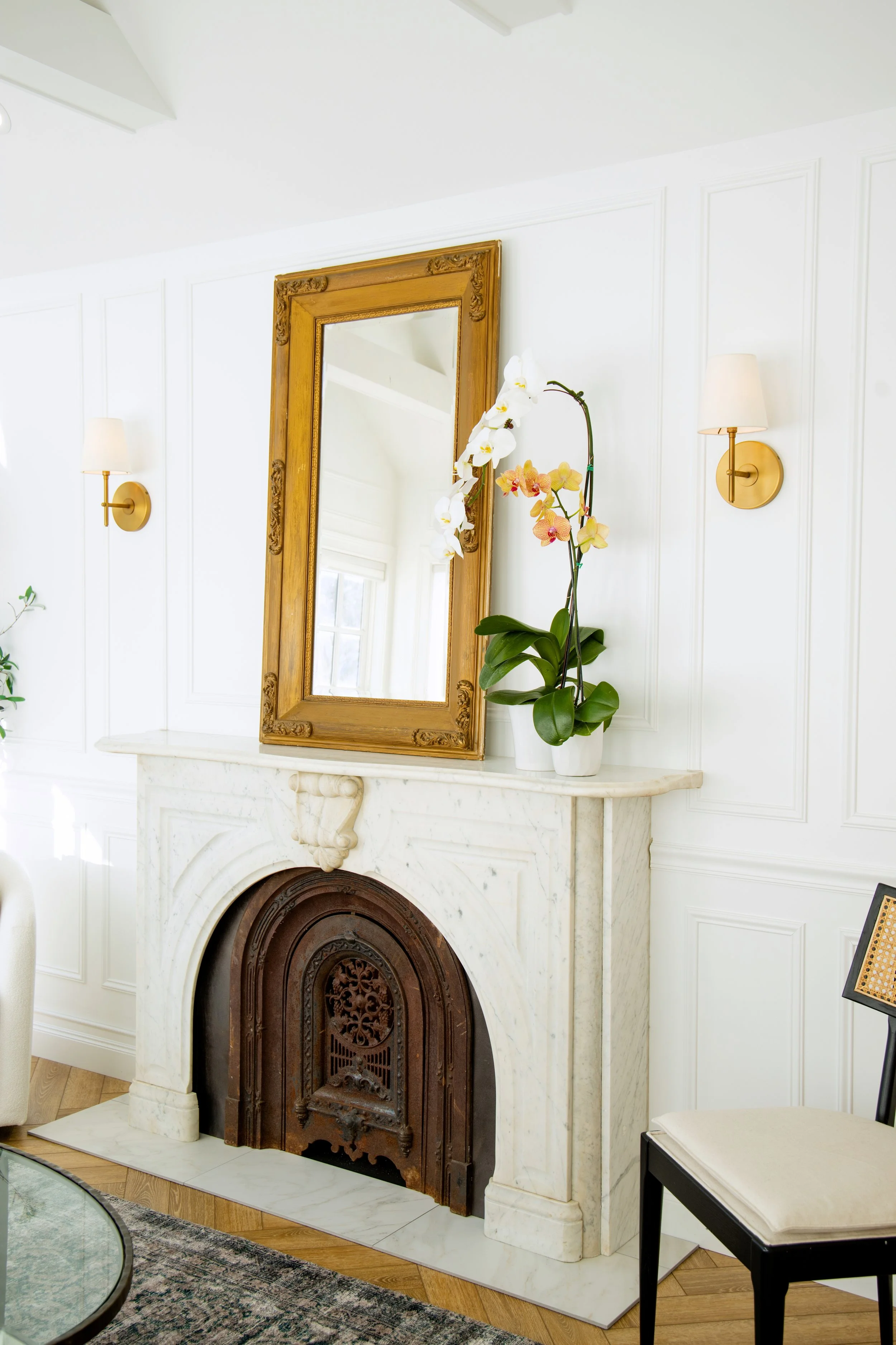 Interior of a room with a white marble fireplace, a mirror with a gold frame above it, and two wall-mounted lamps with white shades. There is a potted orchid with white and yellow flowers on the mantel and a black chair with a beige cushion to the ri