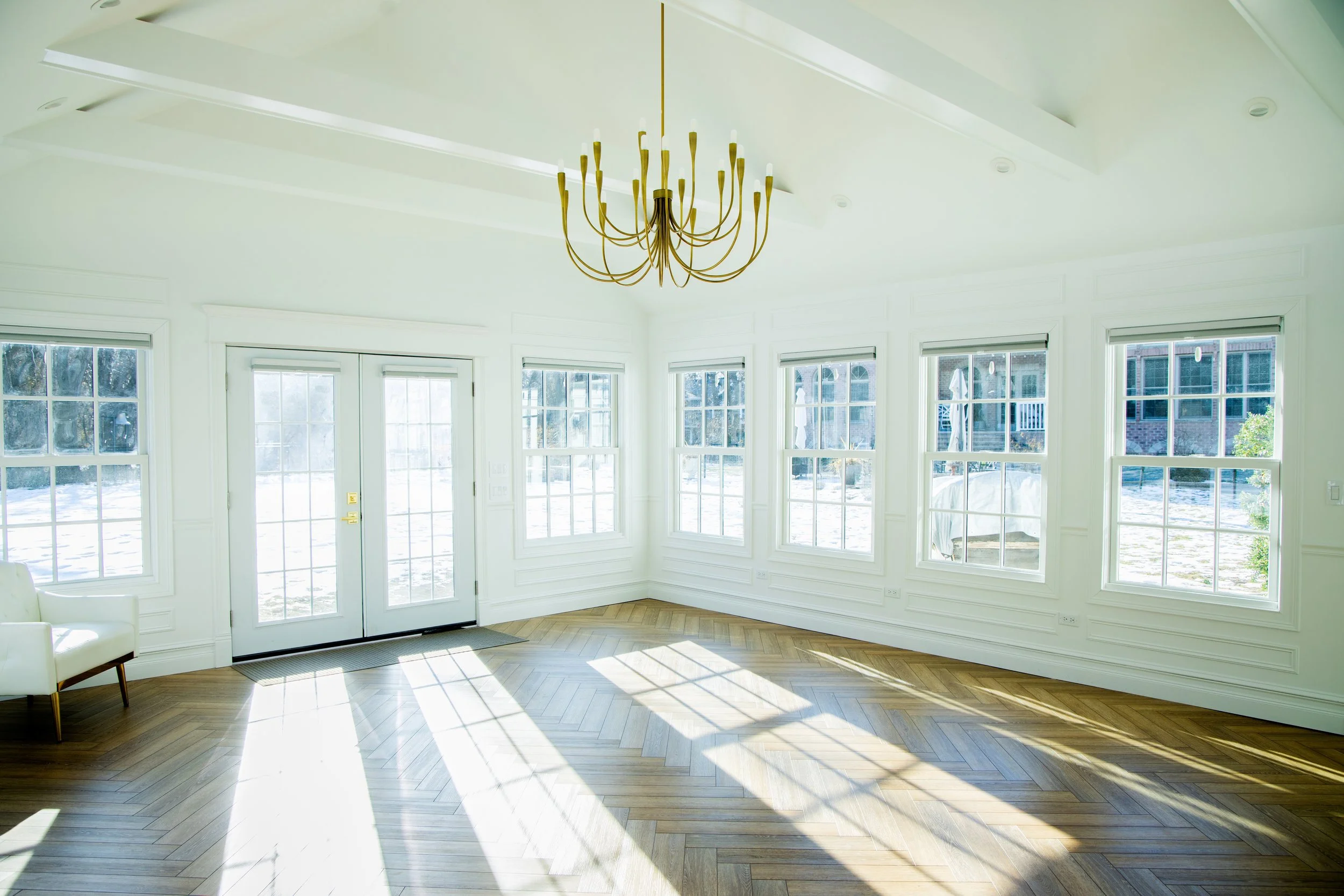 Bright, empty sunlit living room with large windows, a white armchair, a gold chandelier, and hardwood floors.