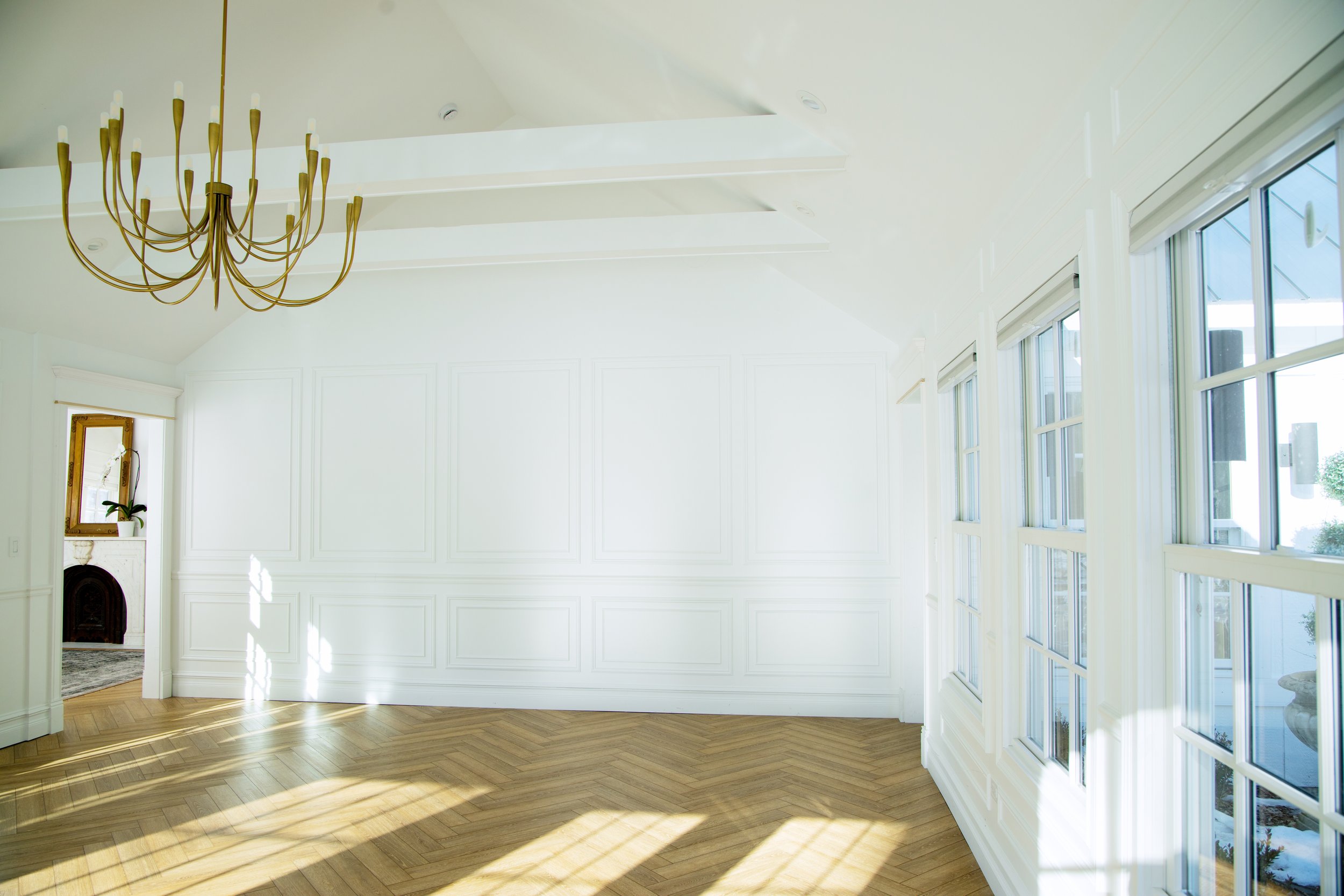 Empty room with white walls, large windows, wooden floor, chandelier, and a small mirror with a plant in the background.