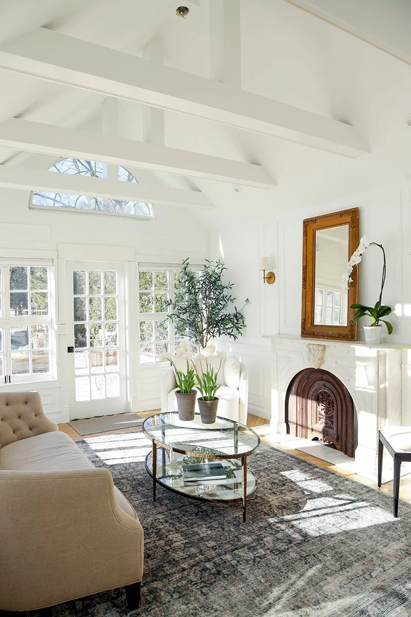 Bright living room with white walls, large windows, a beige sofa, a glass coffee table, an ornate fireplace, potted plants, and a wooden-framed mirror.