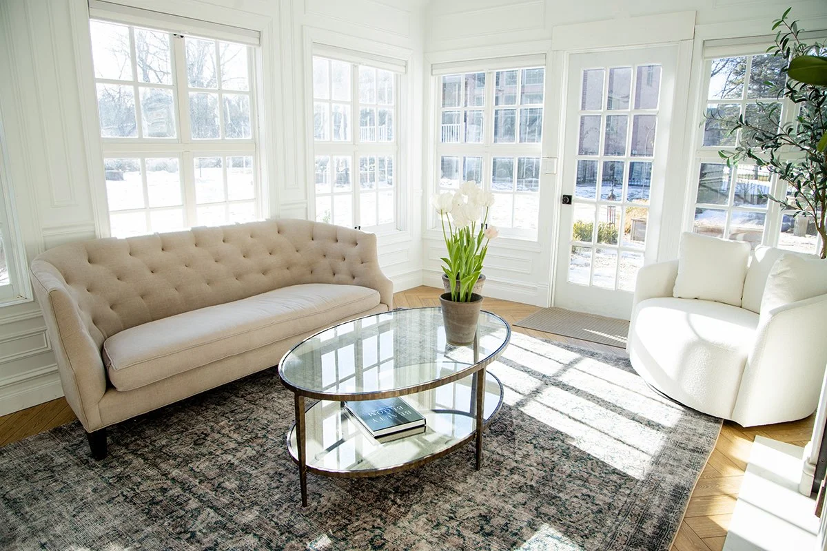 Bright living room with large windows, beige tufted sofa, white armchair, glass coffee table with a flowerpot of white tulips, patterned rug, and green plant in corner.