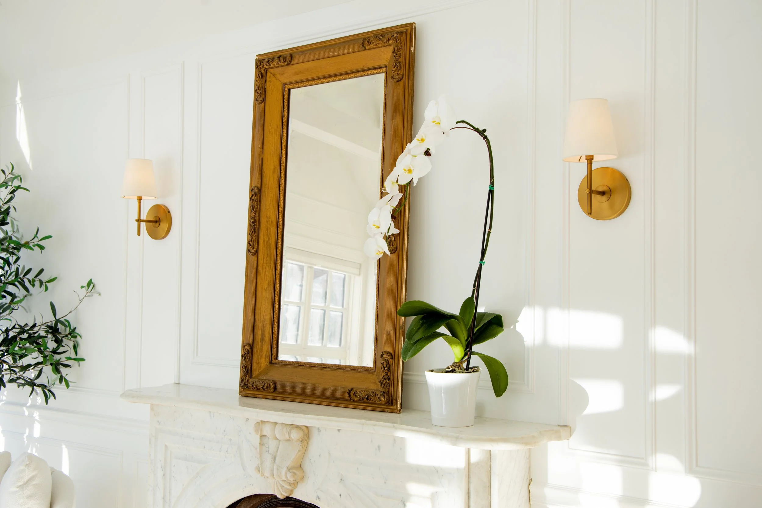 A white orchid in a white pot placed on a marble fireplace mantel, with a wooden framed mirror and two wall-mounted lamps in a bright room with white paneled walls.