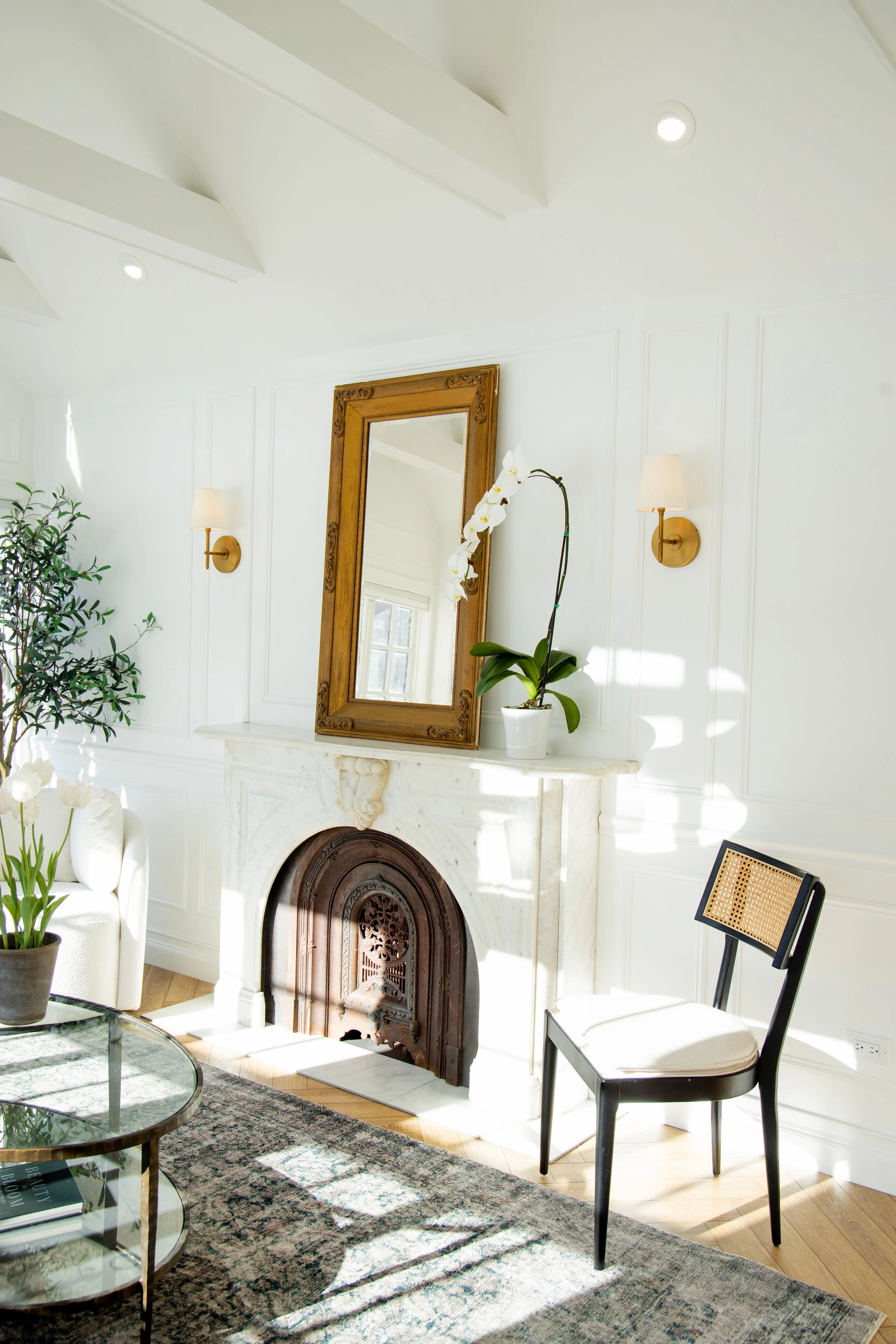 Living room with white walls, a marble fireplace, a large mirror, a potted orchid and additional plants, a patterned area rug, a glass coffee table, and a black wooden chair with a woven back.