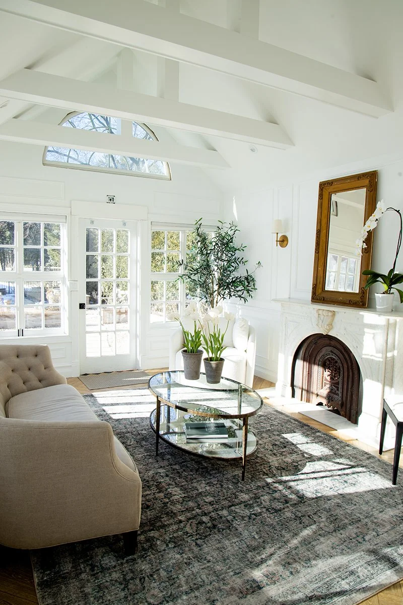 Bright living room with white walls, high ceiling with beams, large windows, a beige couch, a glass coffee table, a fireplace, and potted plants.