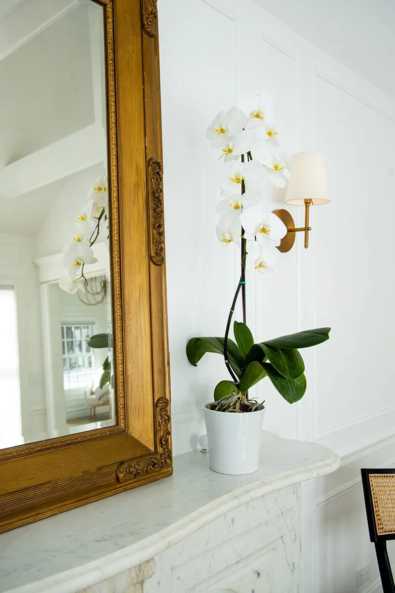 A white orchid in a white pot placed on a marble surface next to a large mirror with a carved wooden frame. A wall with a sconce light fixture is visible in the background.