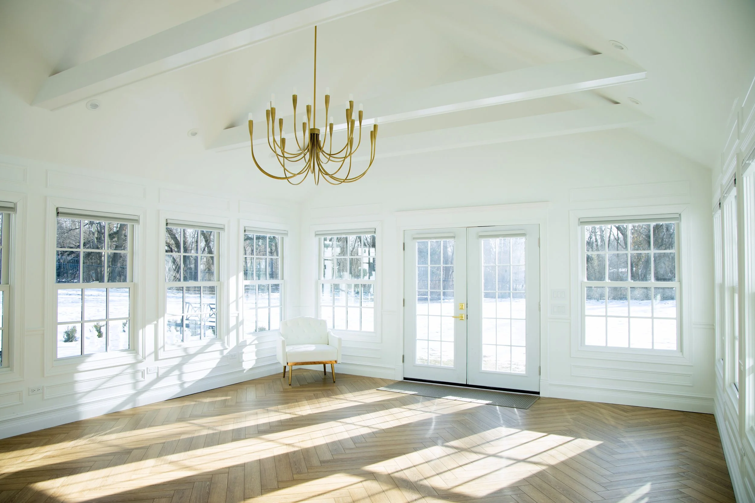 Bright living room with white walls, large windows letting in natural light, a gold chandelier, a white armchair, and view of outdoor snow-covered landscape.