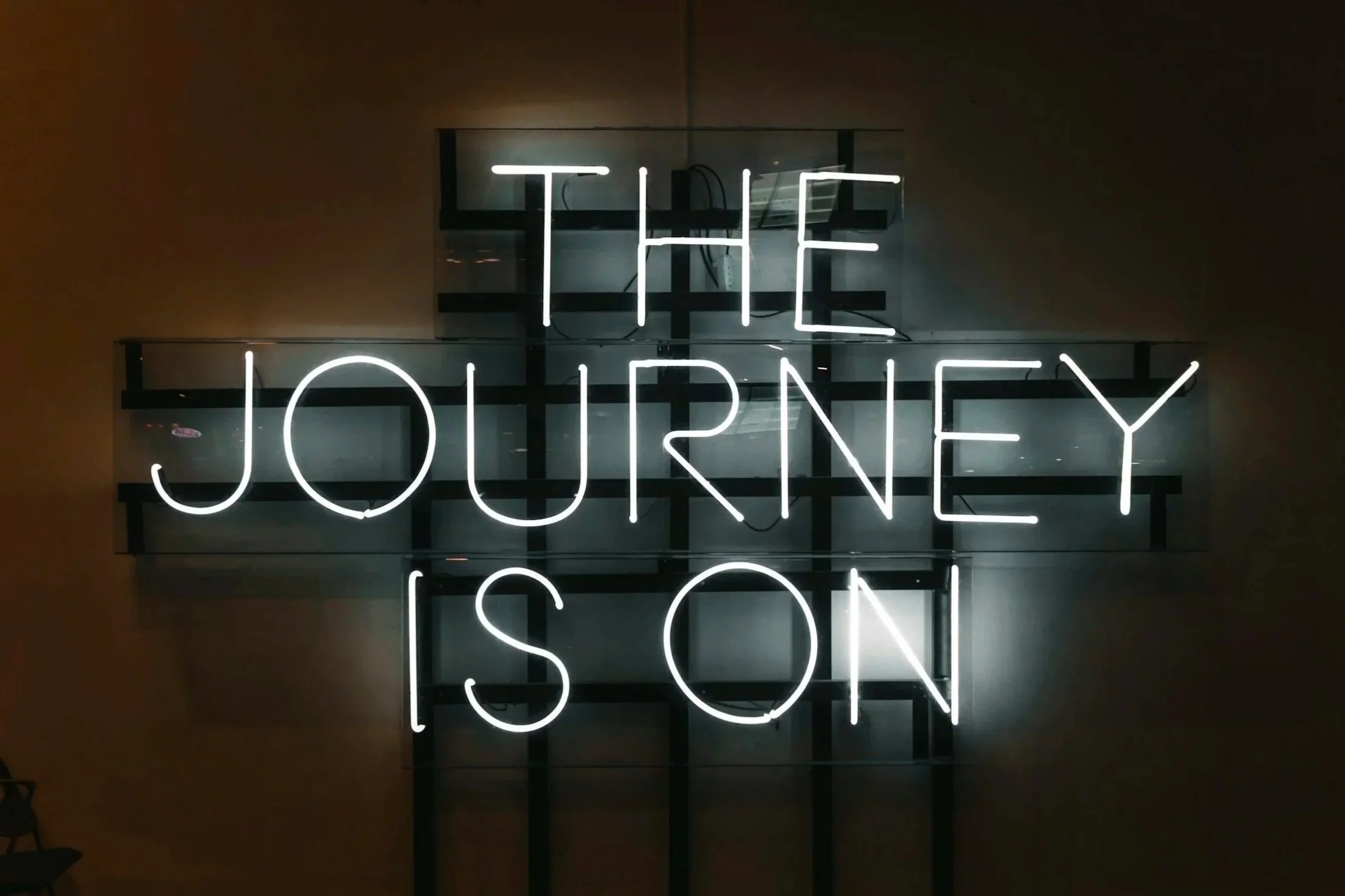 Neon sign with the words “The journey is on” glowing against a dark wall.