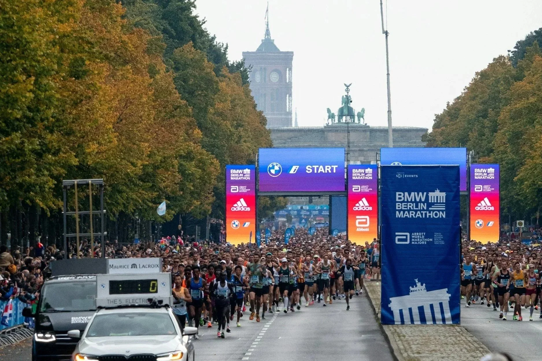 Large group of runners starting the Berlin Marathon at the official start line with race banners and autumn trees lining the road.