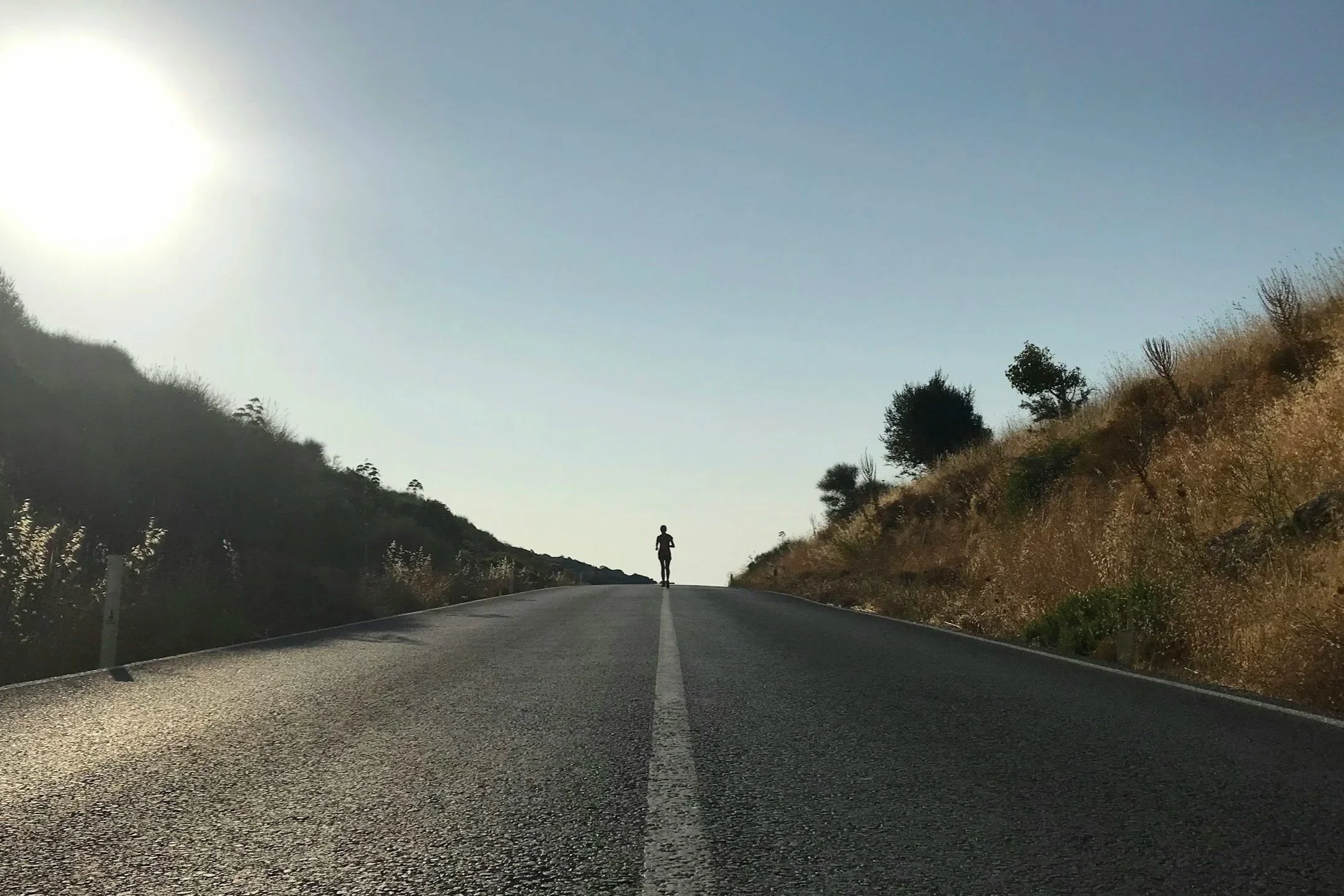 A lone runner silhouetted on an empty road at sunrise, running uphill between dry hills.