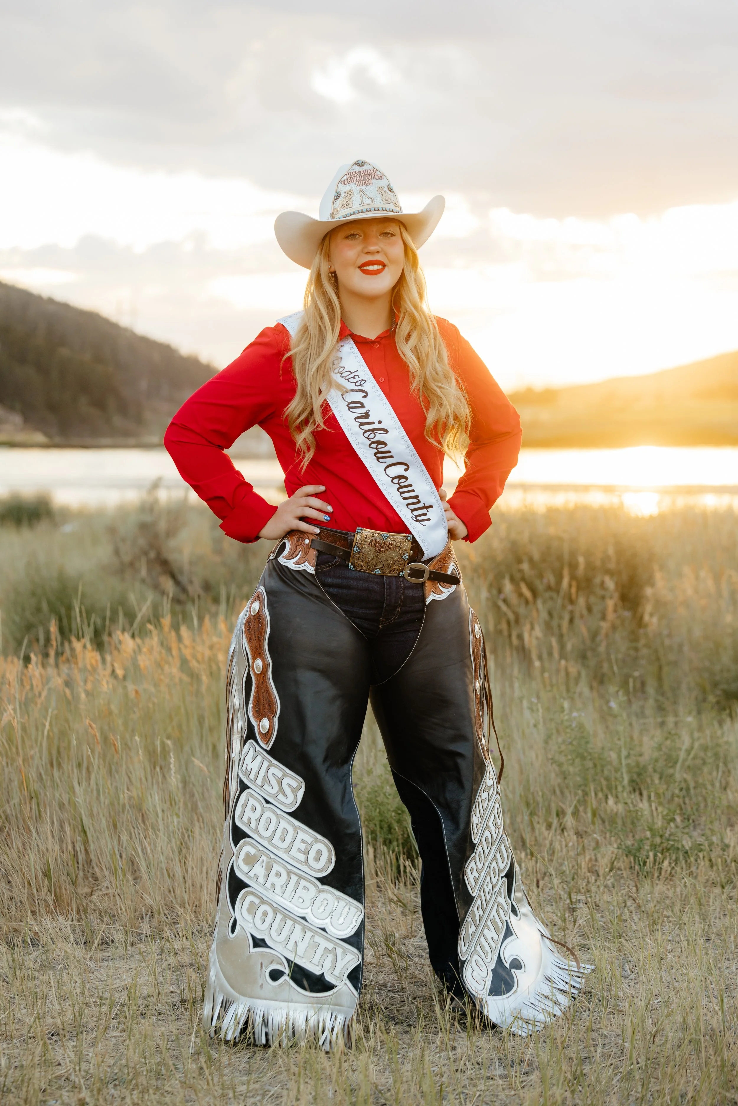 A young woman in Rodeo Queen attire standing confidently on a rodeo arena at sunset, wearing a white cowboy hat, black button-up shirt, large decorative belt, and wide-leg chaps with 'Miss Rodeo Caribou County' printed on them.