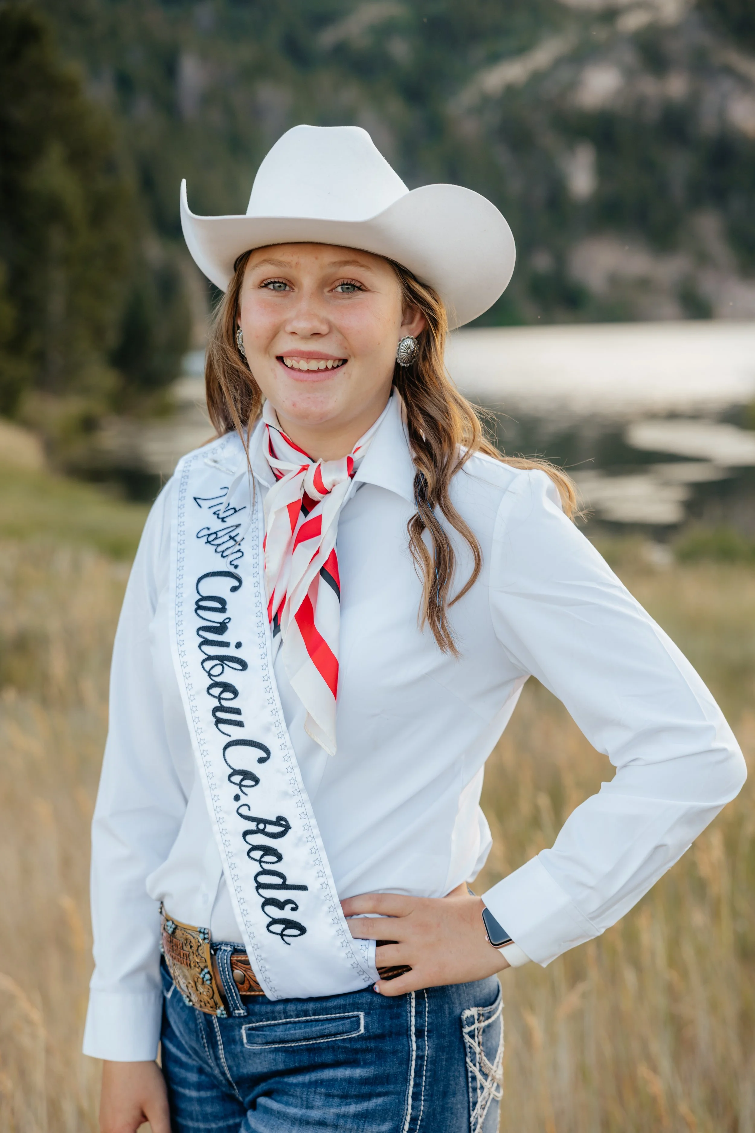 Young woman wearing a white cowboy hat, red plaid shirt, and blue jeans, standing in a rodeo arena with a sash that reads "2nd Runner Up, Auburnt, 2025." She has blonde hair, freckles, and a slight smile.