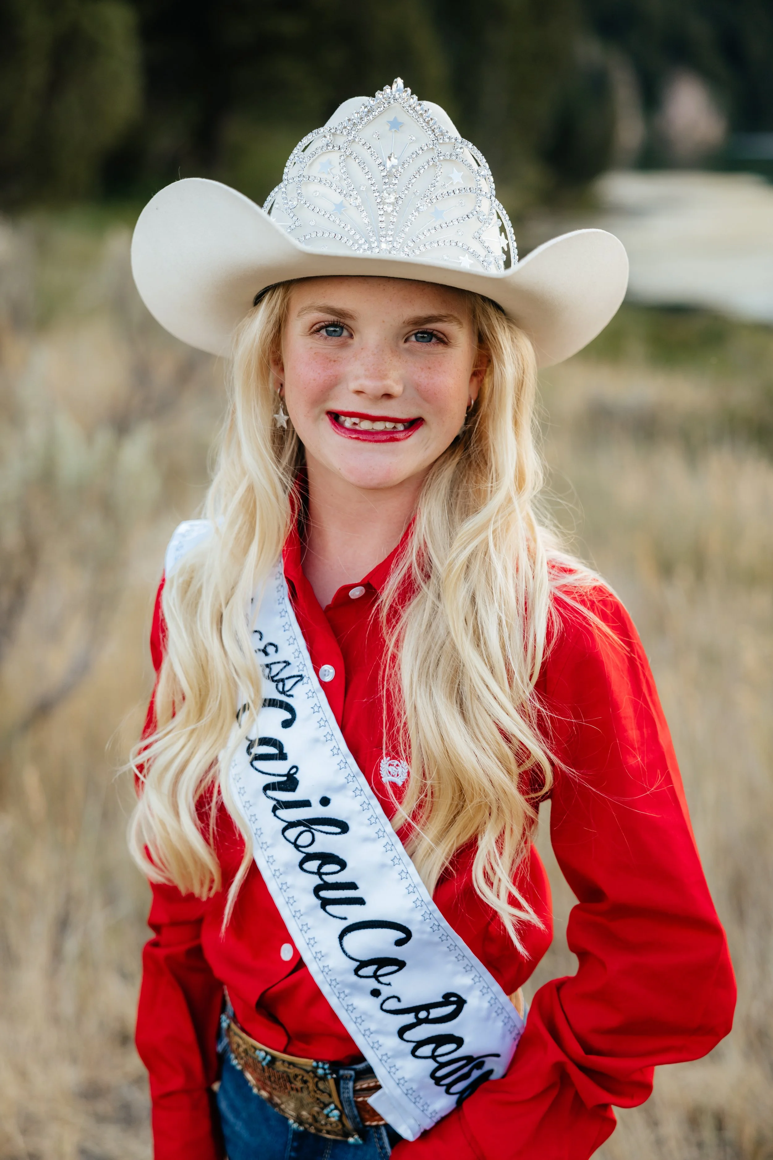 A young girl wearing a black cowboy hat with a silver design, a sash that reads 'Princess' from Caribou County, and a gray and purple long-sleeve shirt, standing on a dirt rodeo arena with a background of a rodeo arena and distant hills.