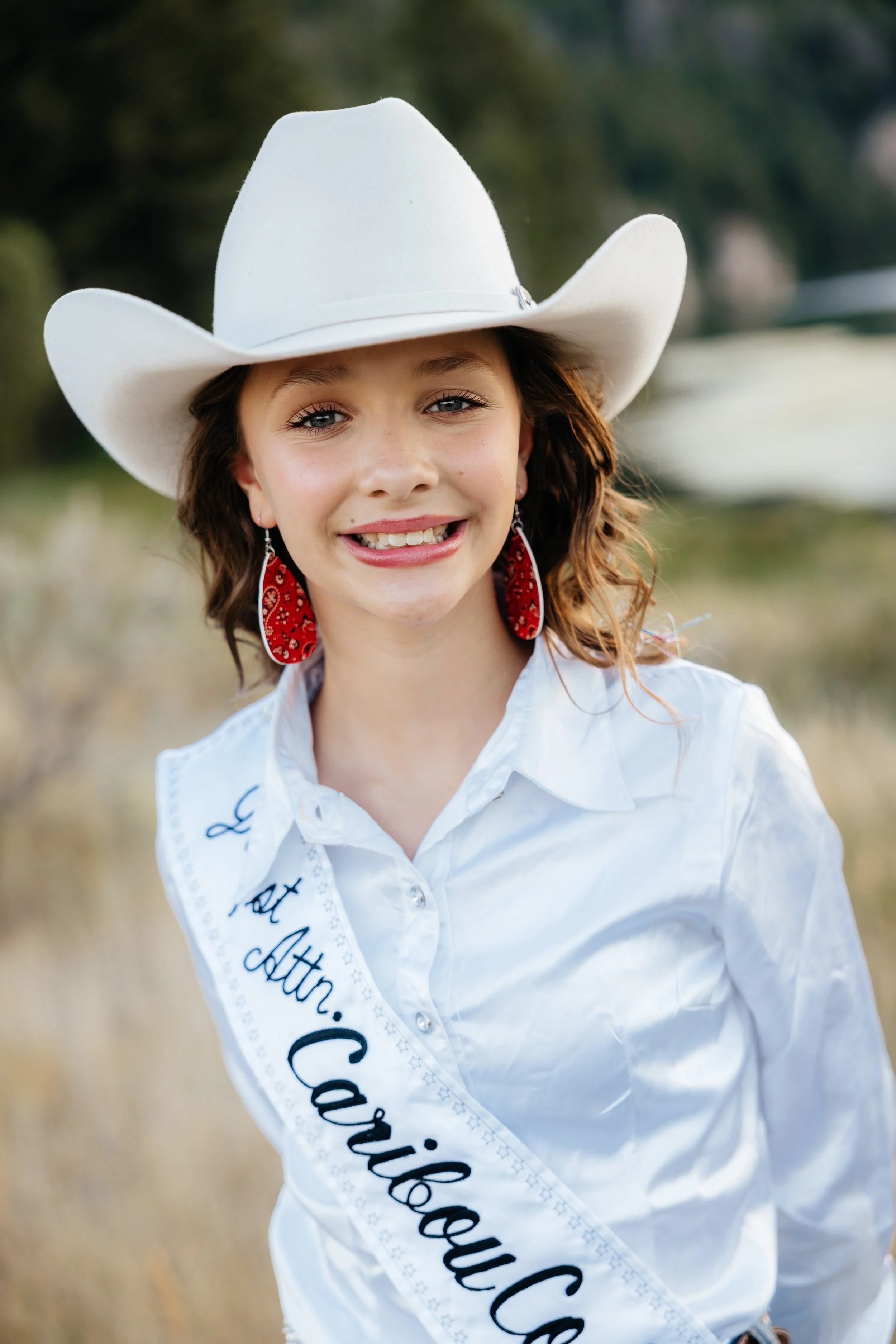 A young girl dressed in pink lace western attire, wearing a white cowboy hat and a sash that reads 'All Around 2025', standing on a dirt rodeo arena with a blue fence and announcer booth in the background.