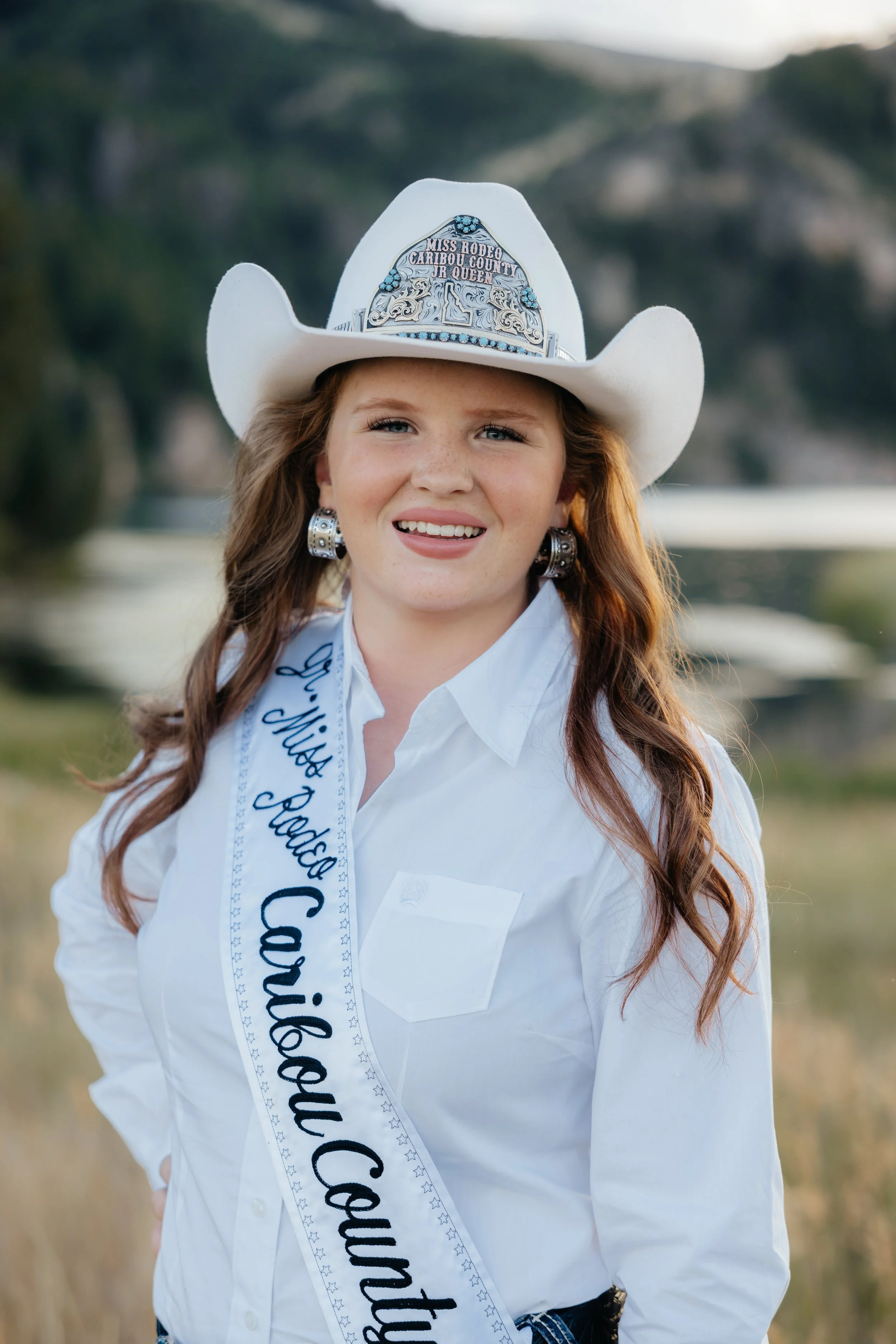 Young girl wearing a cowboy hat, pink Western attire, and a sash that reads 'Jr. Queen' standing on a dirt rodeo arena.