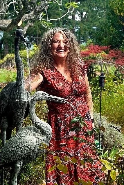 A woman with curly hair smiling and standing outdoors in a garden, next to sculptures of herons.