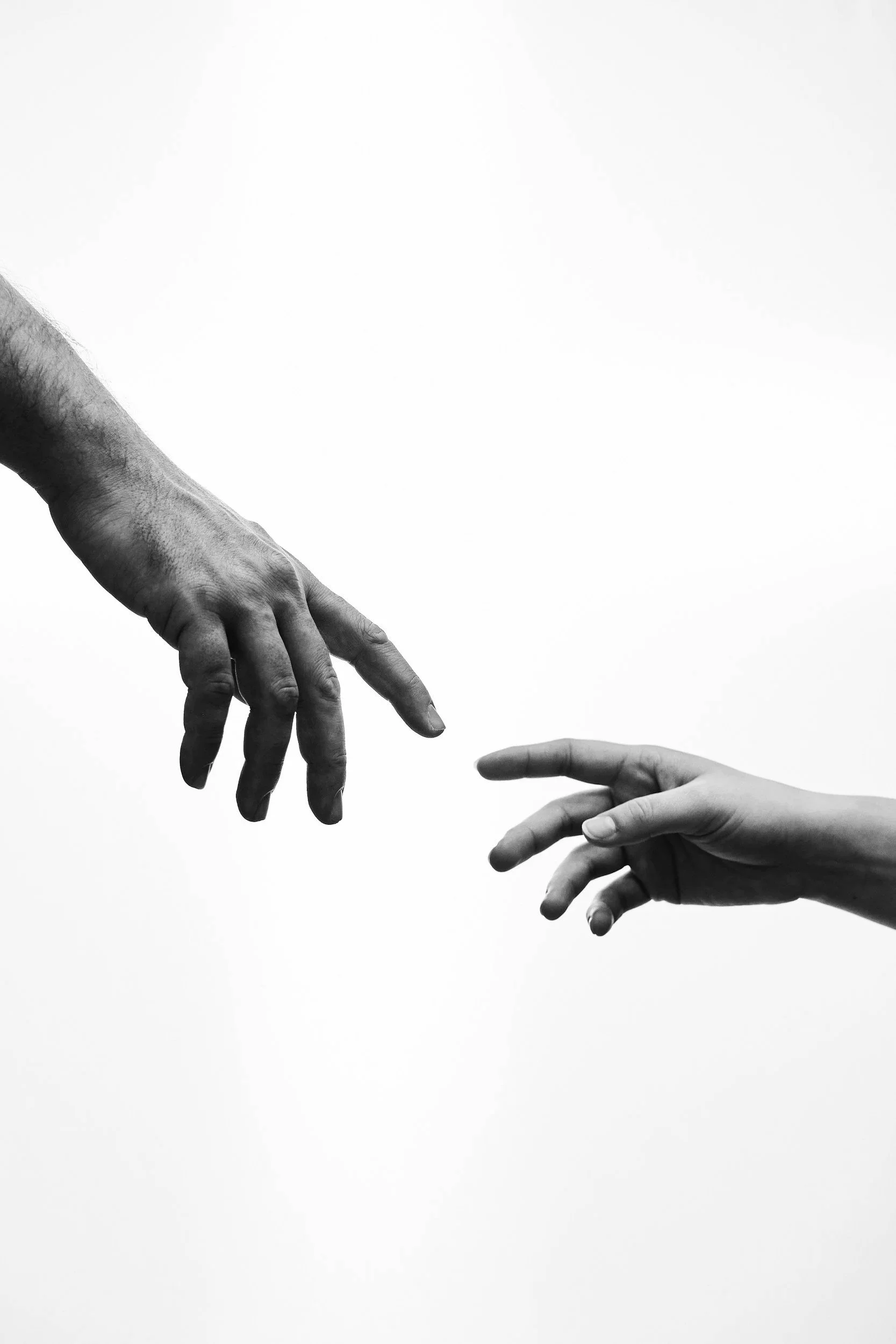 Black and white photo of two hands reaching towards each other, one from the left and one from the right, with fingers almost touching.