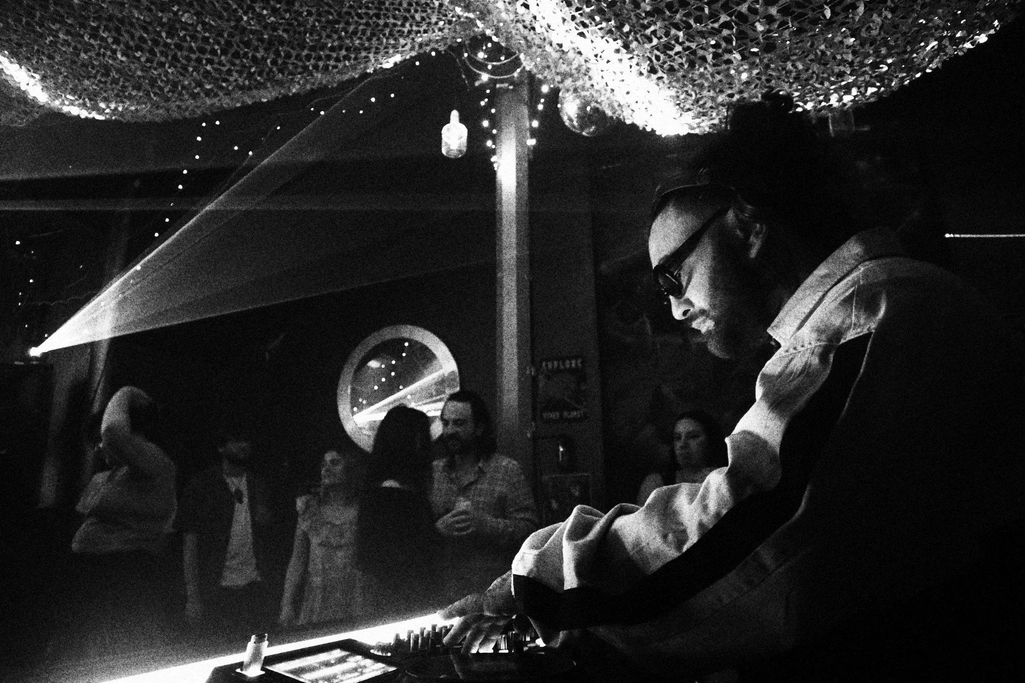 A DJ wearing sunglasses and a white shirt mixing music in a dark nightclub with people dancing in the background and decorative lighting overhead.