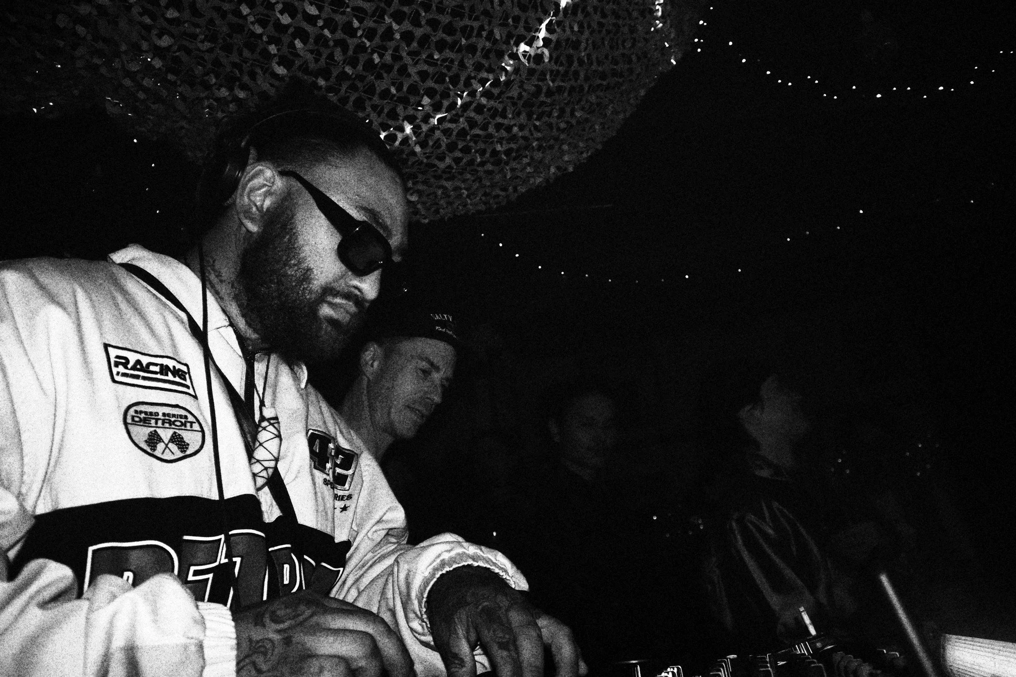 Three people with serious expressions are in a dark room with decorative string lights and a textured ceiling, focused on DJ equipment in front of them.