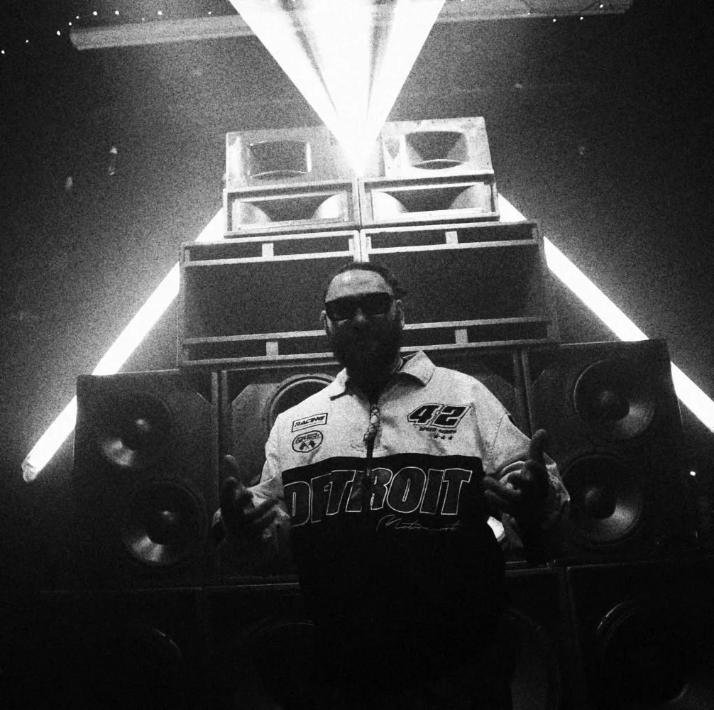 A person wearing sunglasses and a racing jacket with 'Detroit' on it, standing in front of a large speaker setup with neon lights and a triangular light projection above.