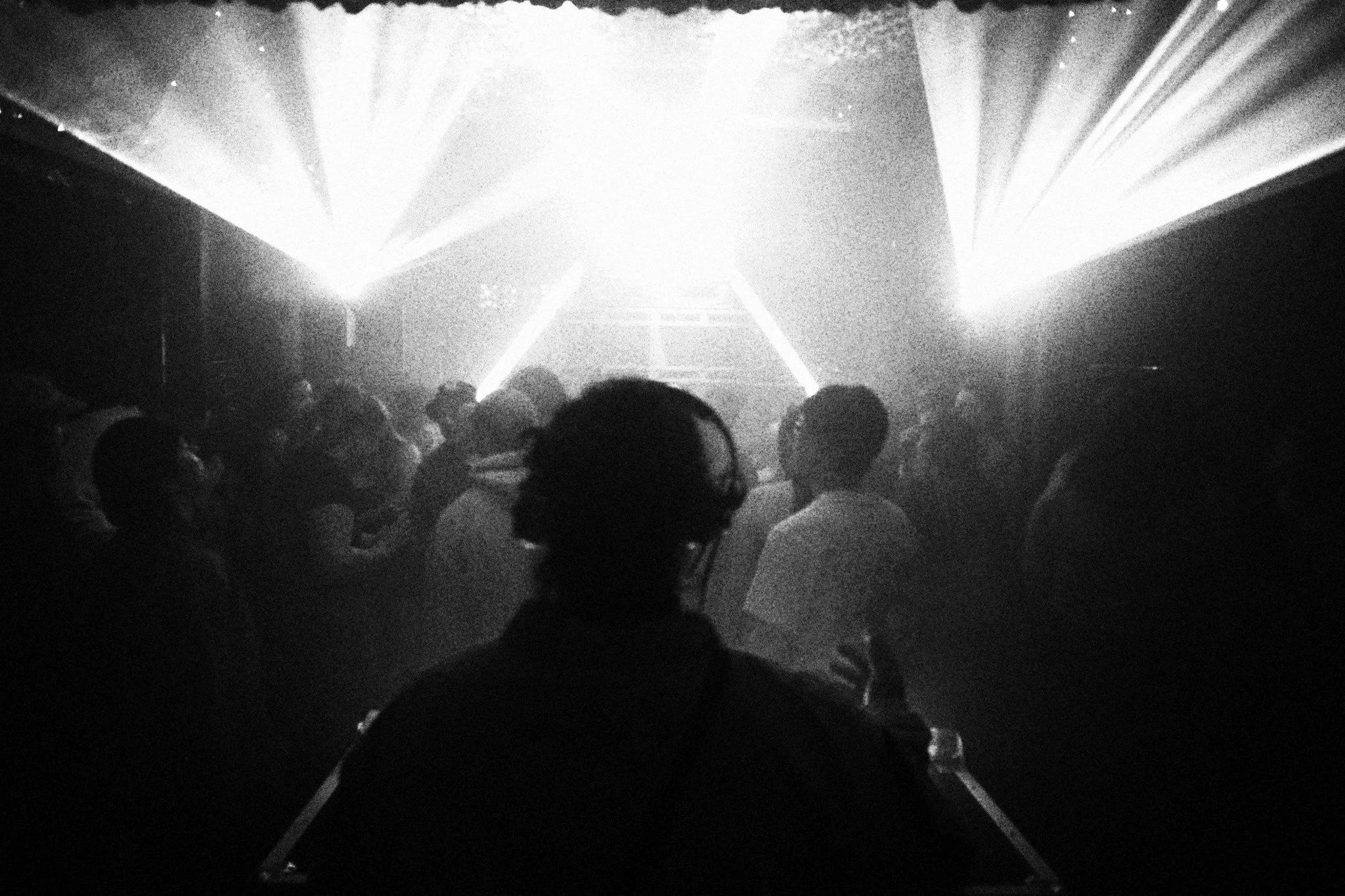 A black and white photo of a nightclub or concert with silhouettes of people dancing and bright stage lights shining in the background.
