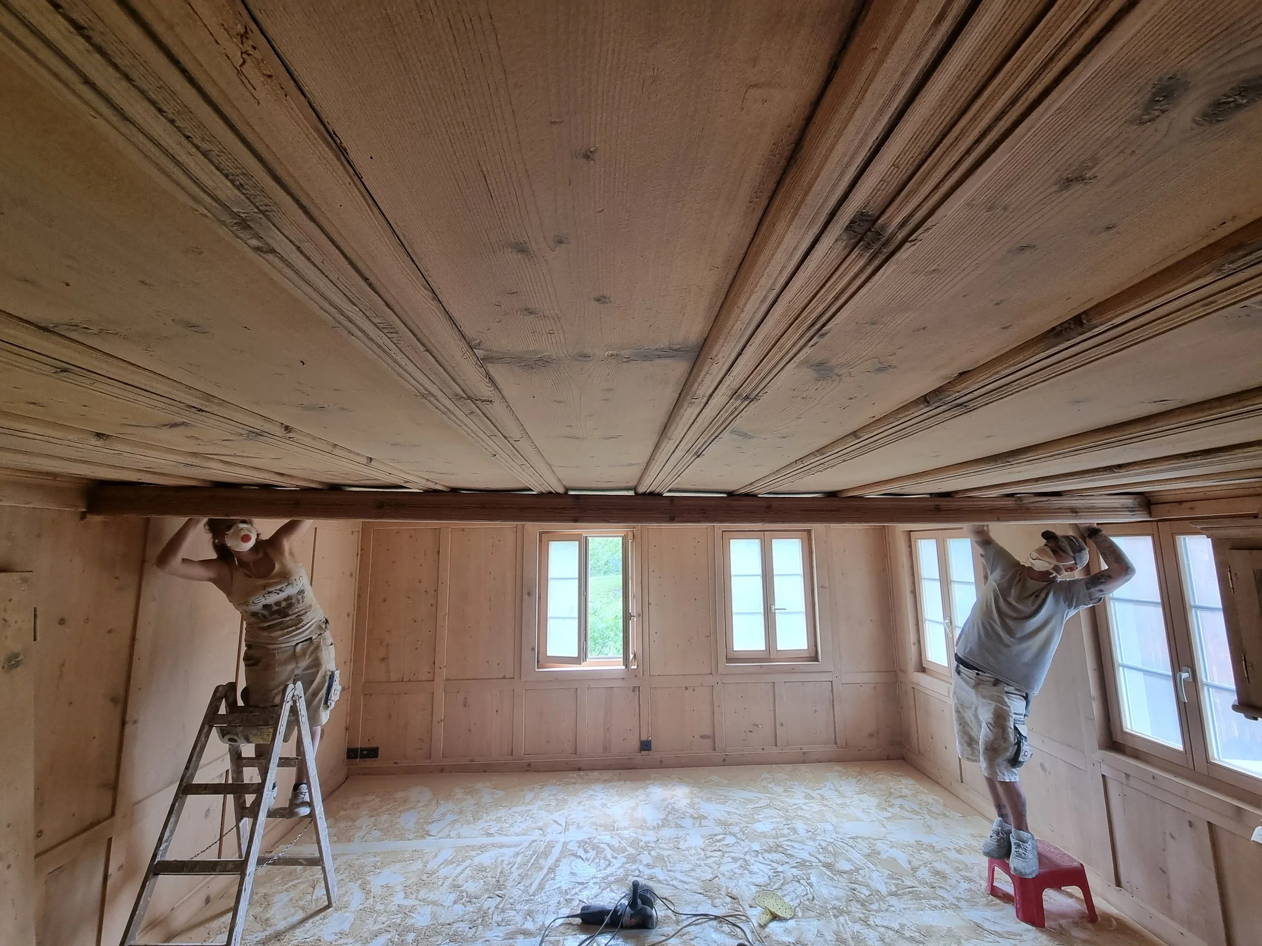Zwei Menschen streichen eine Dachgeschossdecke in einem Raum mit Holzverkleidung. Eine Person steht auf einer Leiter auf der linken Seite, die andere person steht auf einem kleinen Hocker auf der rechten Seite.
