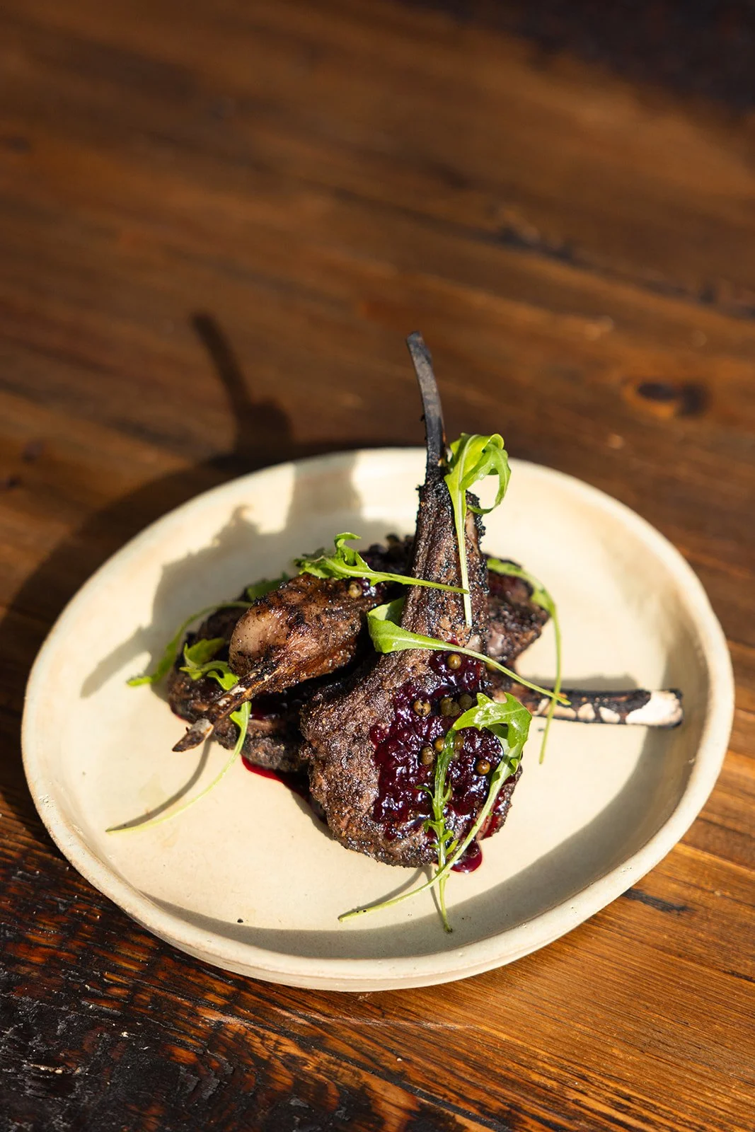 A plate of grilled lamb chops garnished with microgreens and served with a berry sauce on a wooden table.