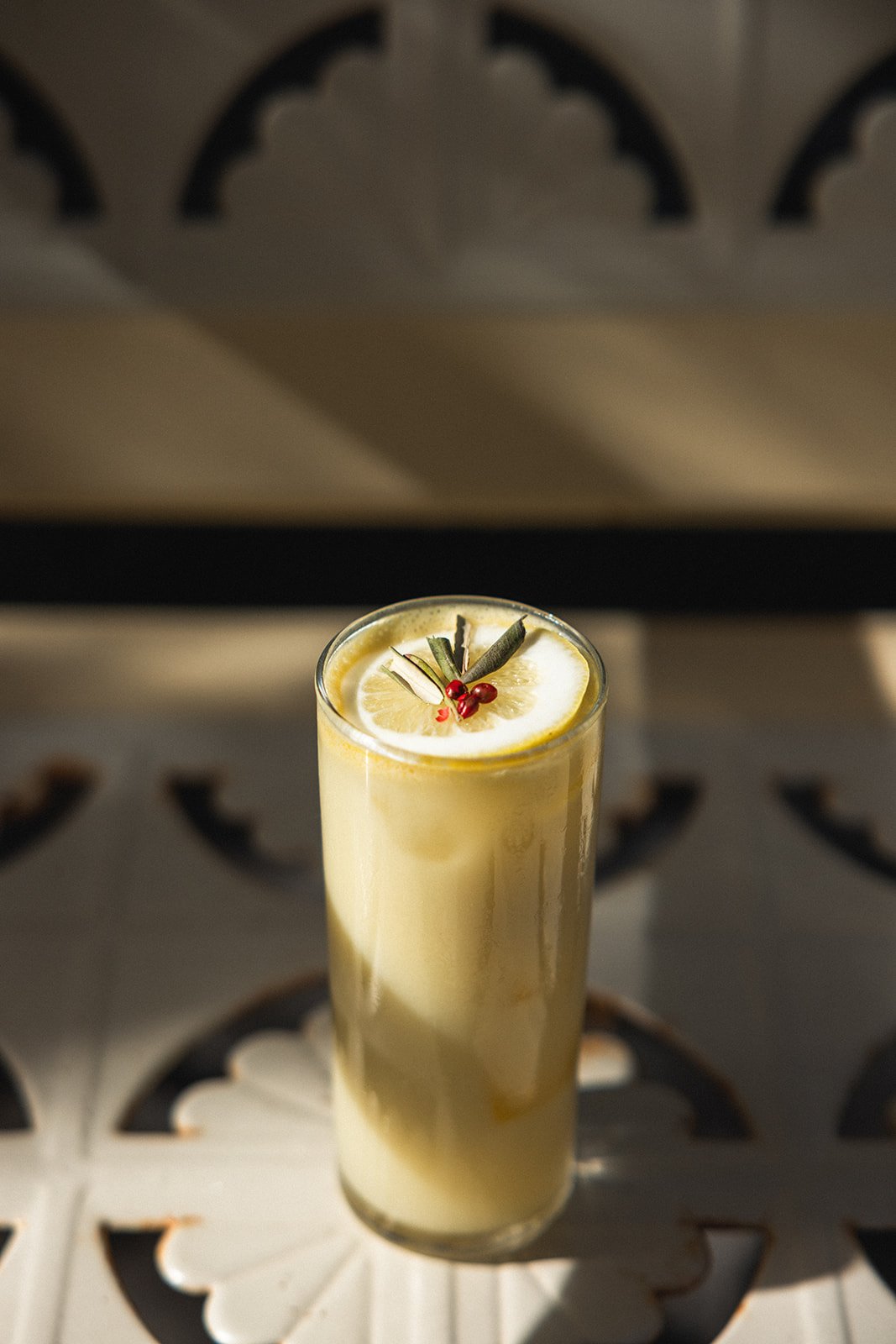 A tall glass of lemon cocktail garnished with a lemon slice, herbal sprigs, and red berries on top, placed on a decorative white surface with a patterned background.