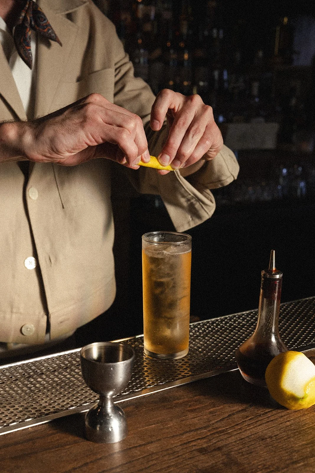 A person wearing a beige blazer and a scarf is squeezing a lemon over a tall glass of yellowish drink with ice at a bar. A metal jigger, a small cruet of dark liquid, and a half lemon are on the bar counter.