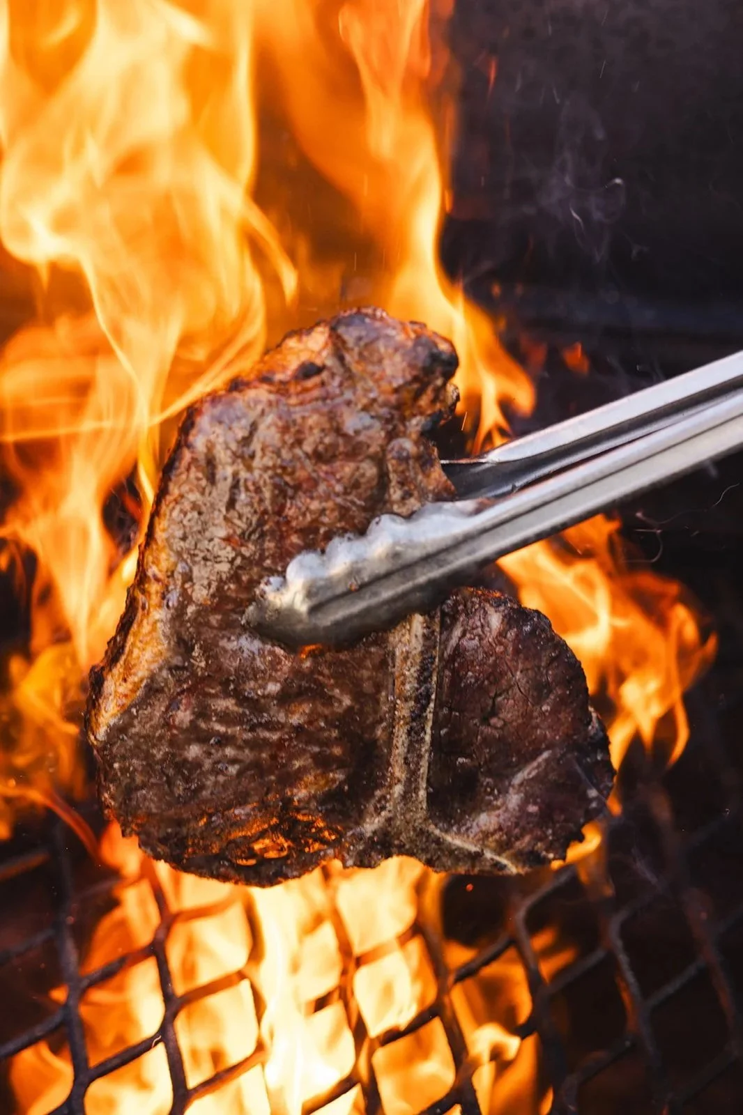 A piece of steak being flipped on a grill over open flames using tongs.