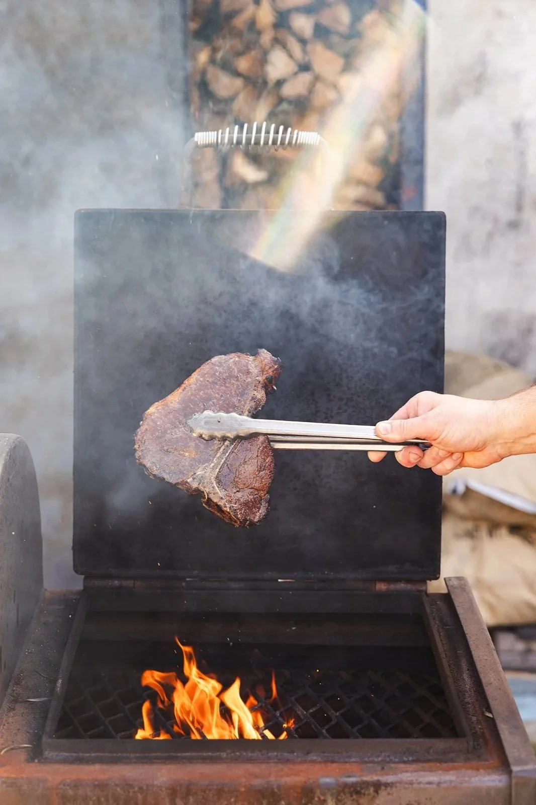 Person using tongs to cook a large piece of meat on an outdoor grill over an open flame.