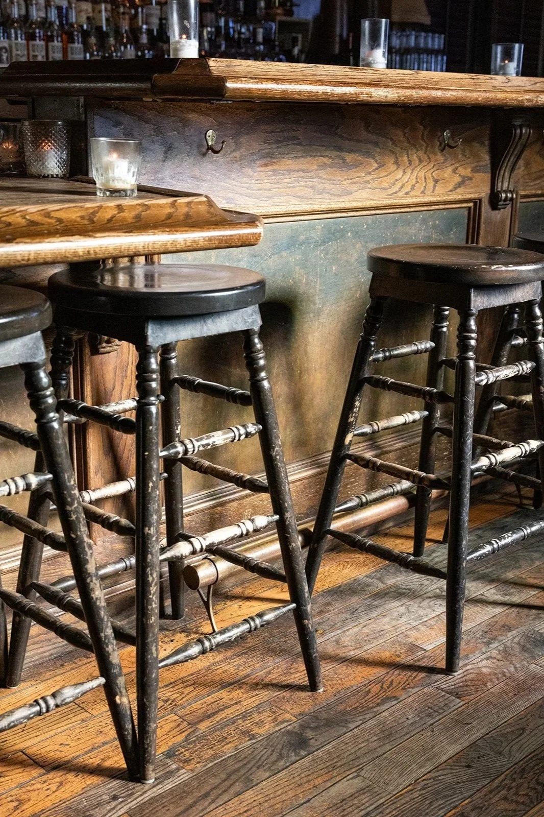 Three black wooden bar stools with worn paint, placed in front of a rustic wooden bar counter in a dimly lit bar or restaurant.