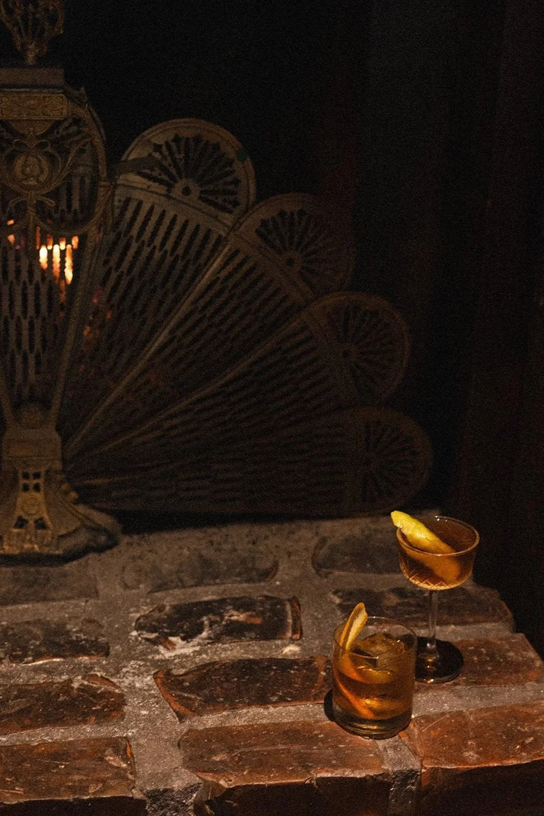 Two cocktails with lemon slices on a brick bar counter, in a dimly lit setting with a large decorative lamp in the background.