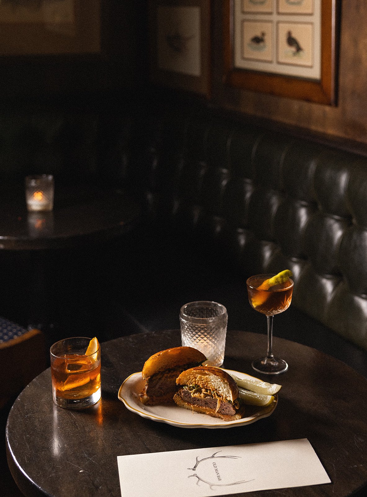A dinner table with a partially eaten beef burger, two pickle spears, and two cocktails garnished with lemon wedges, on a dark wooden table in a dimly lit restaurant.