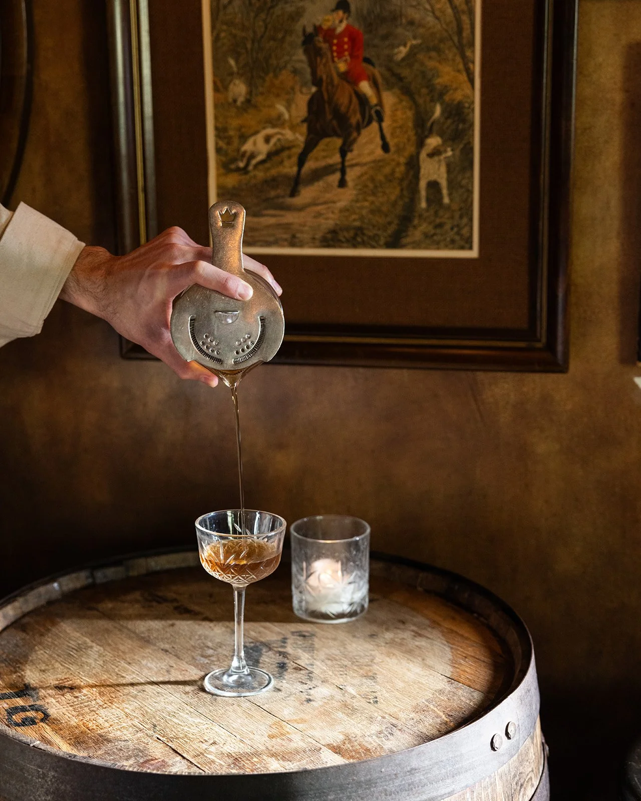 A bartender pouring an amber-colored drink from a metallic bottle into a vintage glass on a wooden barrel, with a lit candle and framed artwork of a man riding a horse in the background.