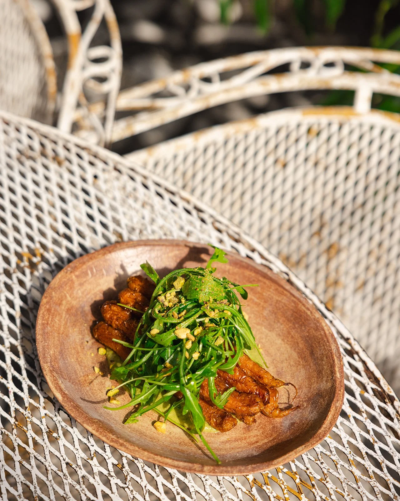 A wooden plate with strips of cooked meat topped with arugula and crushed pistachios, placed on a white metal outdoor table.