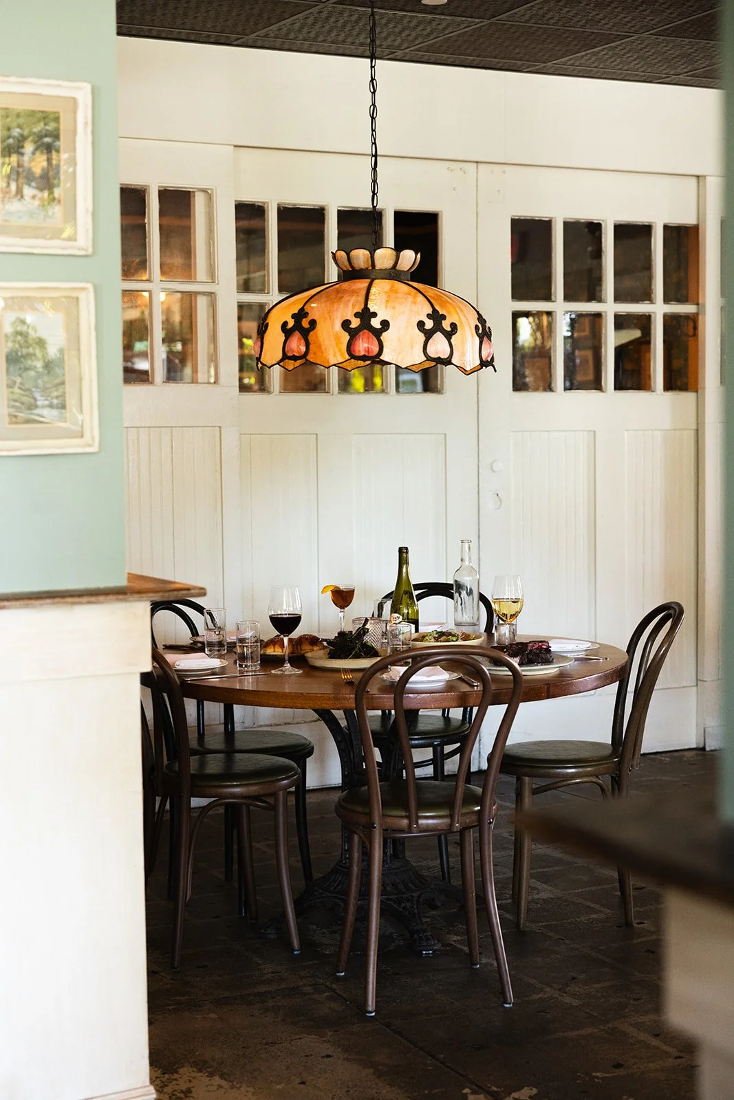 Dining table set with wine glasses, bottles, and food under a Tiffany-style stained glass pendant lamp in a cozy restaurant.