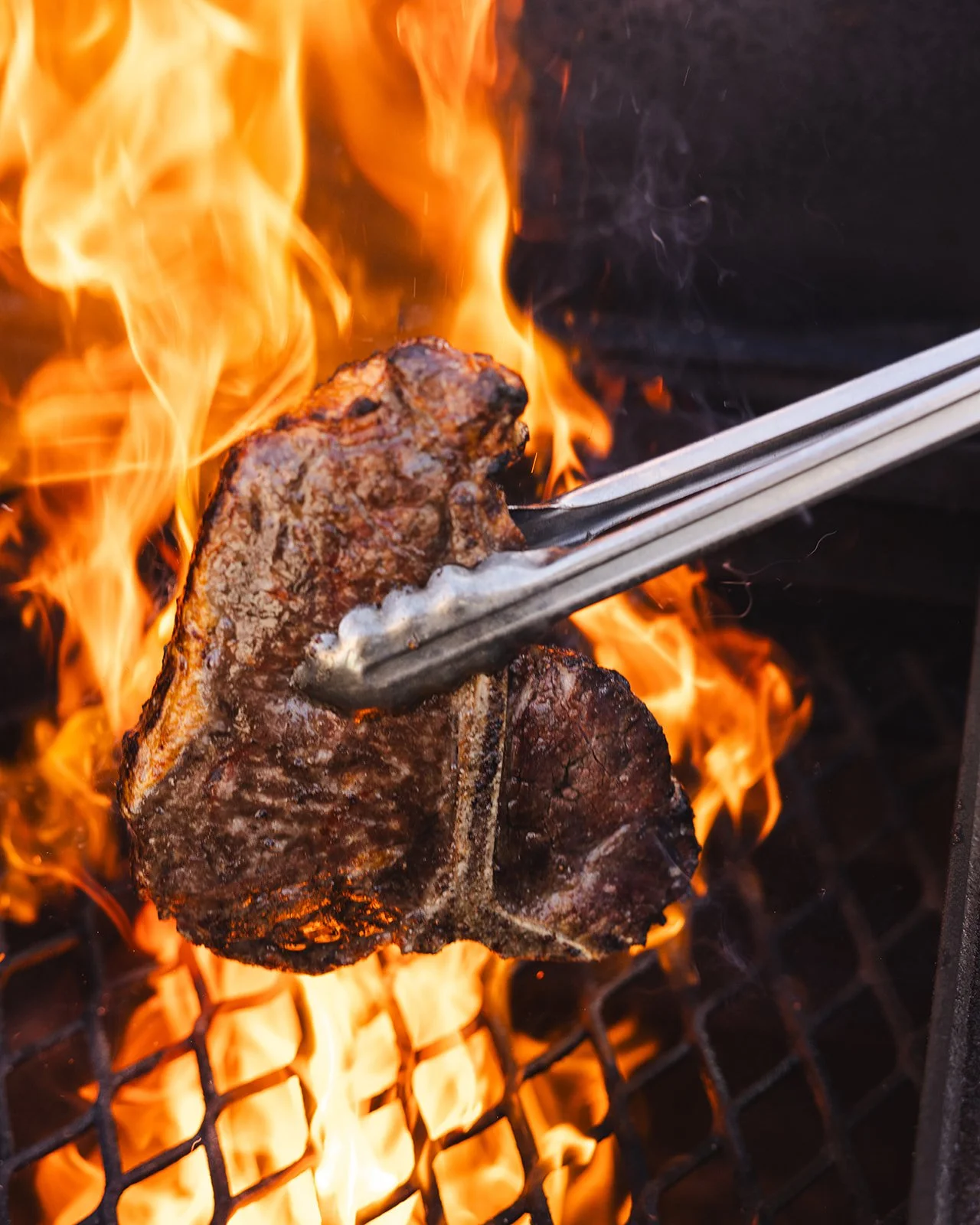 A piece of steak being flipped on a grill over open flames using tongs.