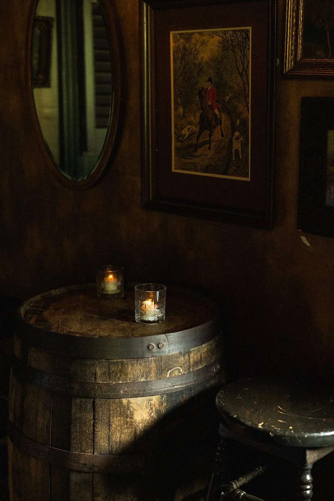 A dimly lit room with candles on a wooden barrel, framed artwork on the wall, and a stool in front of the barrel.