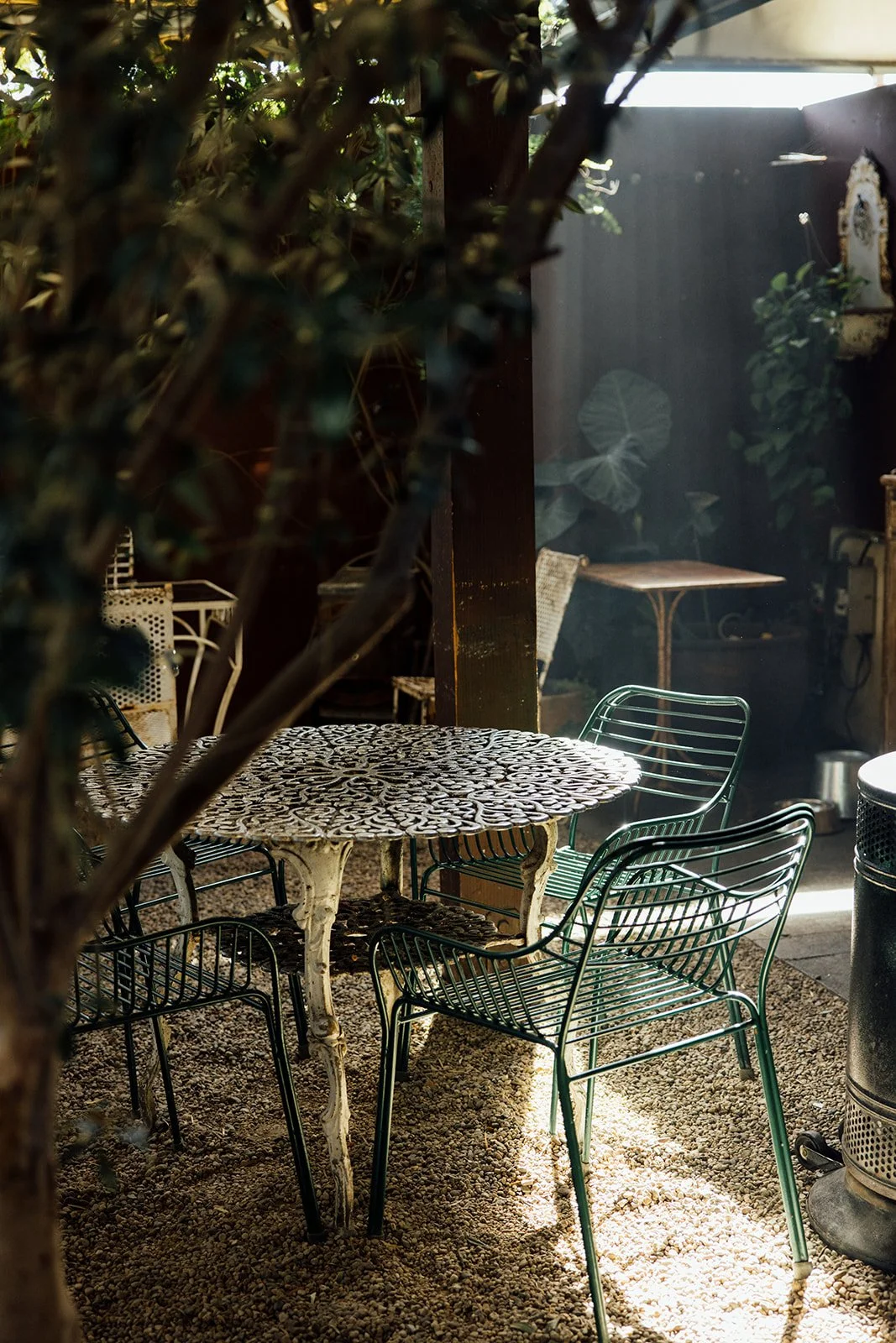 Outdoor patio with a round, ornate metal table and four green wire chairs on a gravel ground, partially shaded by trees, with sunlight filtering through leaves.