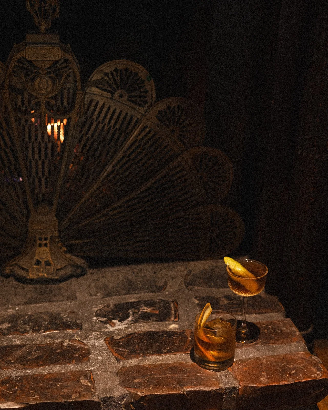 Two cocktails with lemon slices on a brick bar counter, in a dimly lit setting with a large decorative lamp in the background.