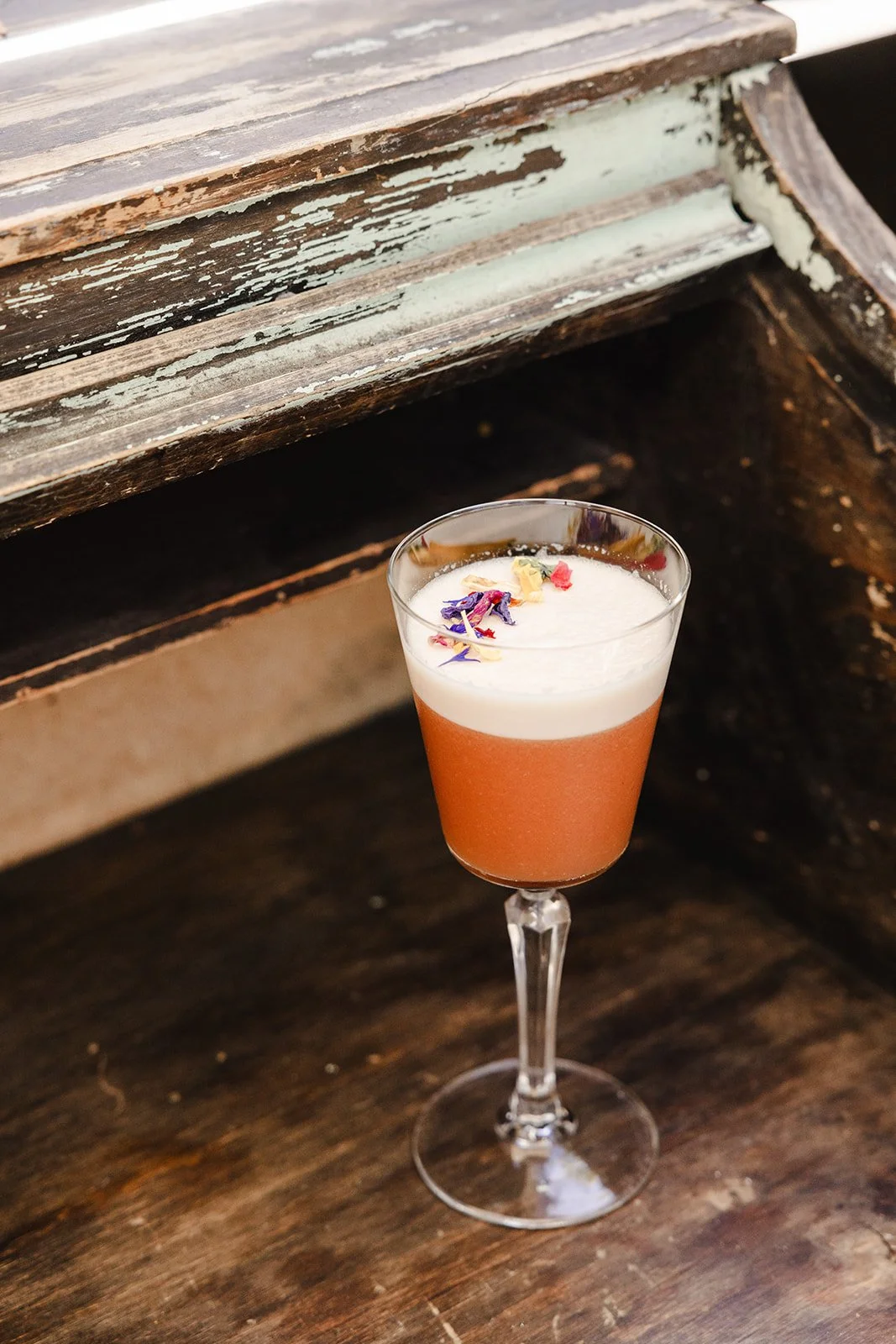 A cocktail in a tall, elegant glass with a frothy white top and colorful flower petals garnishing, placed on a rustic wooden surface.